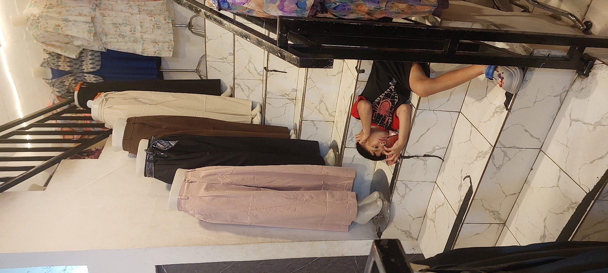 In Jayapura, Indonesia, a young boy in a black t-shirt with a red spider-web design and red sleeves lies on his side on the marble-tiled steps of an indoor staircase. He is holding a mobile phone to his ear with his left hand, while his right hand is bent upwards and touching his ear, suggesting he is talking on the phone or listening intently. The boy is wearing black shorts and a grey shoe with blue accents and a Mickey Mouse charm. The staircase is lined with mannequins displaying women's clothing, including light pink, black, brown, and beige wide-leg pants, as well as a floral dress and a patterned jacket over a blue dress displayed further up. The railings of the staircase are made of dark metal. The overall environment appears to be a clothing store.