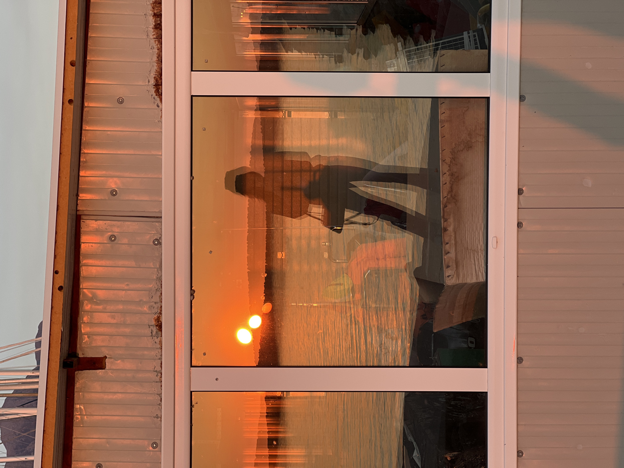 The image, taken in Bar, Montenegro, depicts a sunset reflected in a multi-pane window. Two bright orbs, likely the sun, are visible in the middle pane. Silhouettes of people are superimposed on the sunset reflection in the central pane, along with outlines of objects and structures. The window is set in a corrugated metal wall, secured with visible screws. Parts of what appears to be a boat are visible on the lower left-hand edge of the frame. The light suggests the image was taken during the golden hour of the day.
