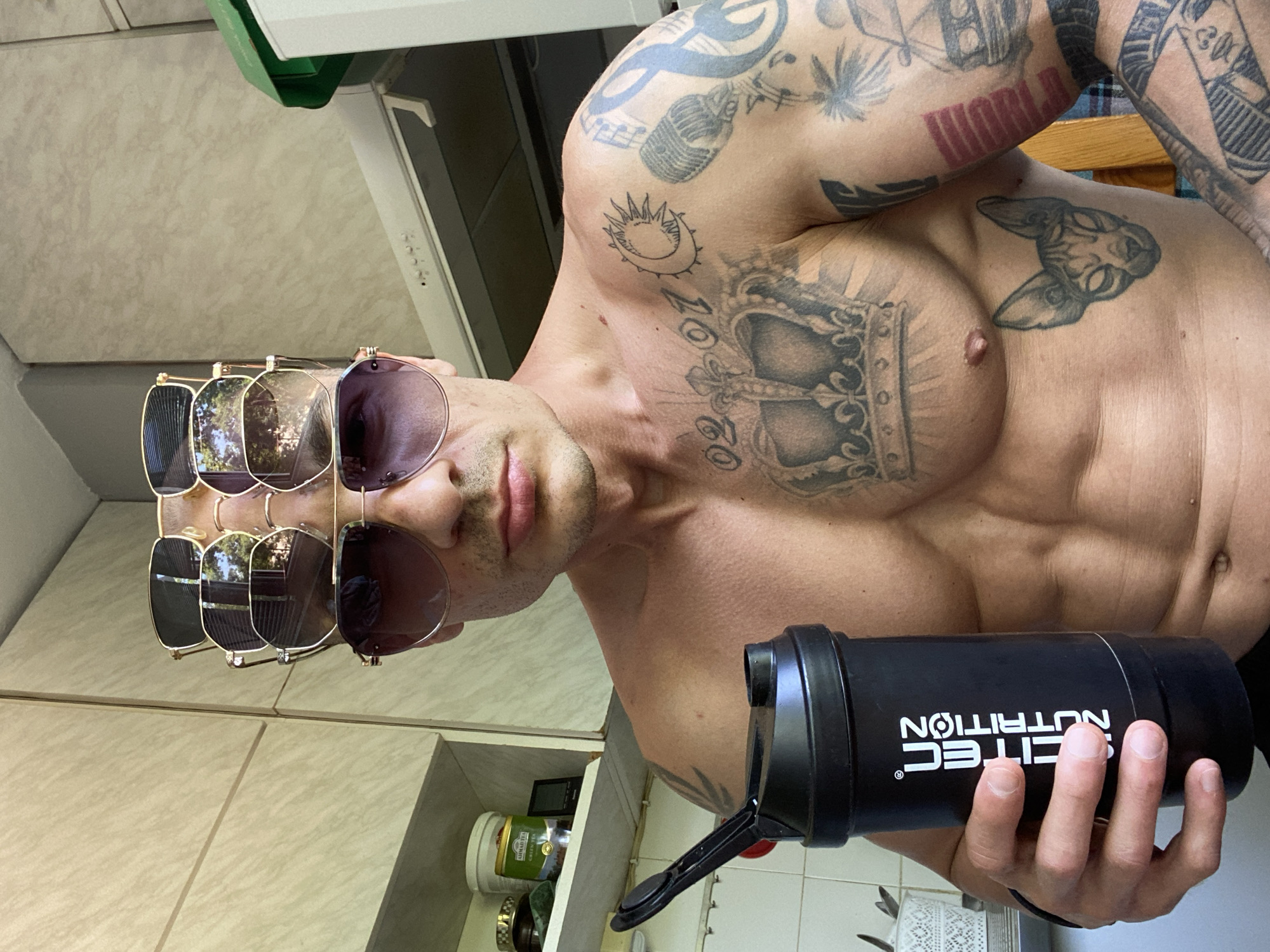 A shirtless man is photographed indoors, holding a black shaker cup labeled "SCITEC NUTRITION." The man has visible tattoos covering his arms, chest, and upper body. He is wearing four pairs of sunglasses stacked on his face. The background includes kitchen cabinets and appliances. The location is likely within a residential building or gym in Pleven, Bulgaria, although specific contextual details to confirm the city are absent.
