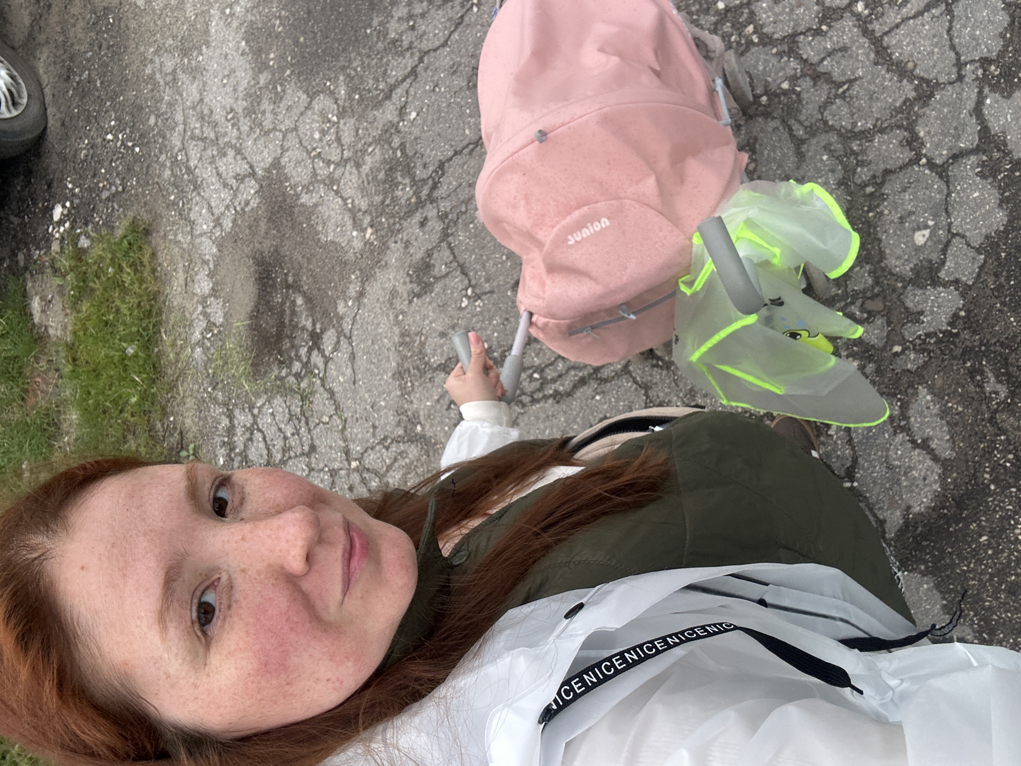 A woman with red hair and freckles is taking a selfie outdoors. She is wearing a green vest over a white shirt with the word "NICE" repeated along the neckline. In the background, there is a pink backpack labeled "JUNION" and a lime green umbrella. The woman is holding the handle of the pink backpack with her left hand, which is attached to a rolling suitcase. The ground is paved with cracked asphalt, and there is some green grass on the left edge of the frame. Given the context of Nizhny Novgorod and the style of the backpack and umbrella, this could be a park or a public space within the city. The photograph gives the impression of a rainy day with the woman traveling or commuting.