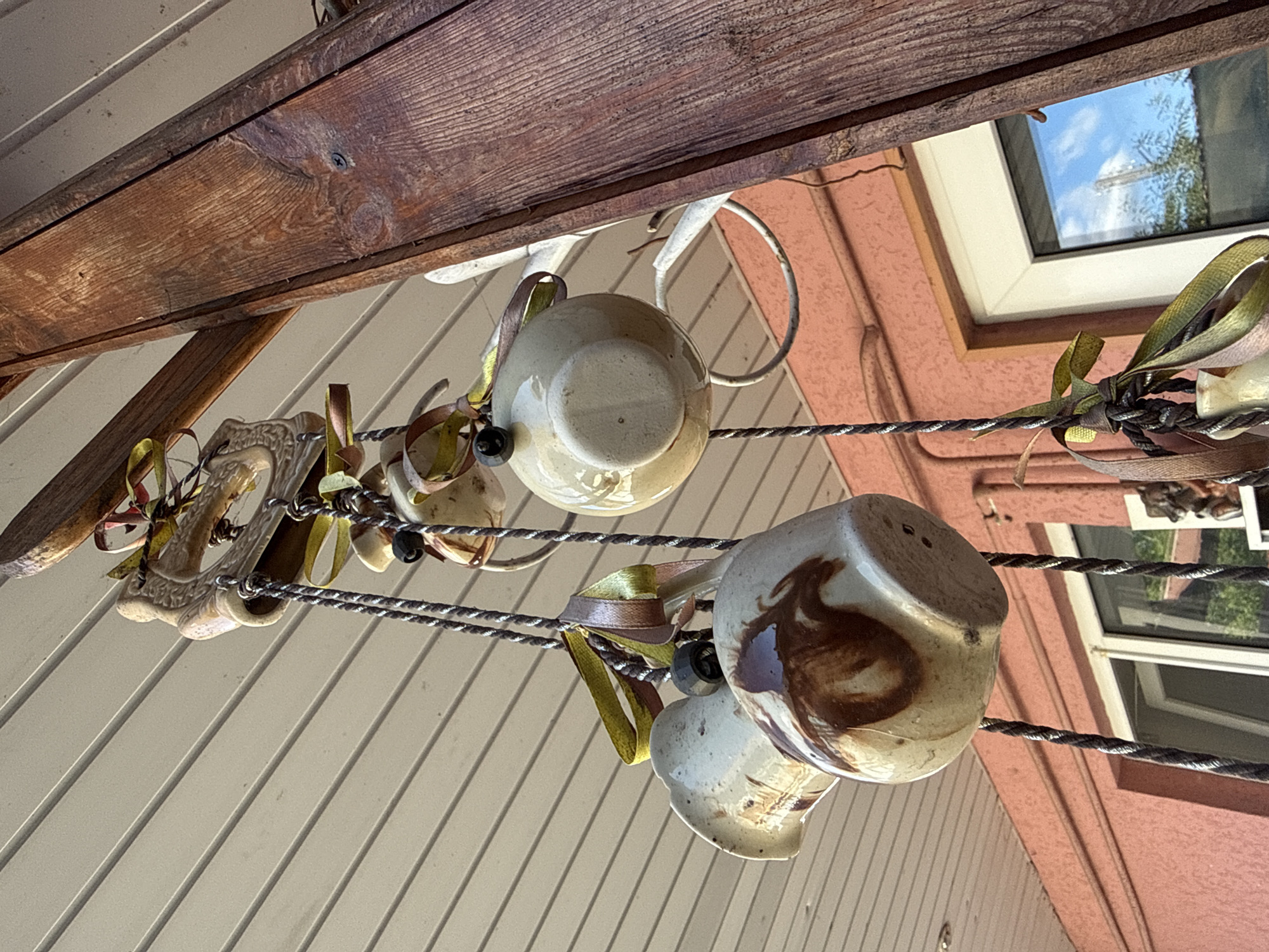 The image captures a close-up, low-angle view of decorative hanging objects suspended from an overhang, likely a porch or eaves, on a sunny day. A thick, weathered wooden beam dominates the right side of the frame, with a visible screw indicating its attachment. Below the beam, a white ceiling with parallel planks extends across the top of the image.
Hanging from ropes are several ceramic decorative elements. One is a pale beige jug with brown marbling, its handle angled upwards. Another is a round, pale beige ceramic sphere. Both items are adorned with colorful ribbons in shades of green, brown, and yellow, tied in bows. A dark, bead-like object is attached to the rope above the jug. At the very top, a decorative ceramic piece, possibly an animal figurine, is visible, also strung with ribbons.
The background shows a salmon-pink wall with a white-framed window. The window reflects a blue sky with clouds and trees. The overall impression is a rustic, outdoor decoration. The location is identified as Okhtyrka, Ukraine.