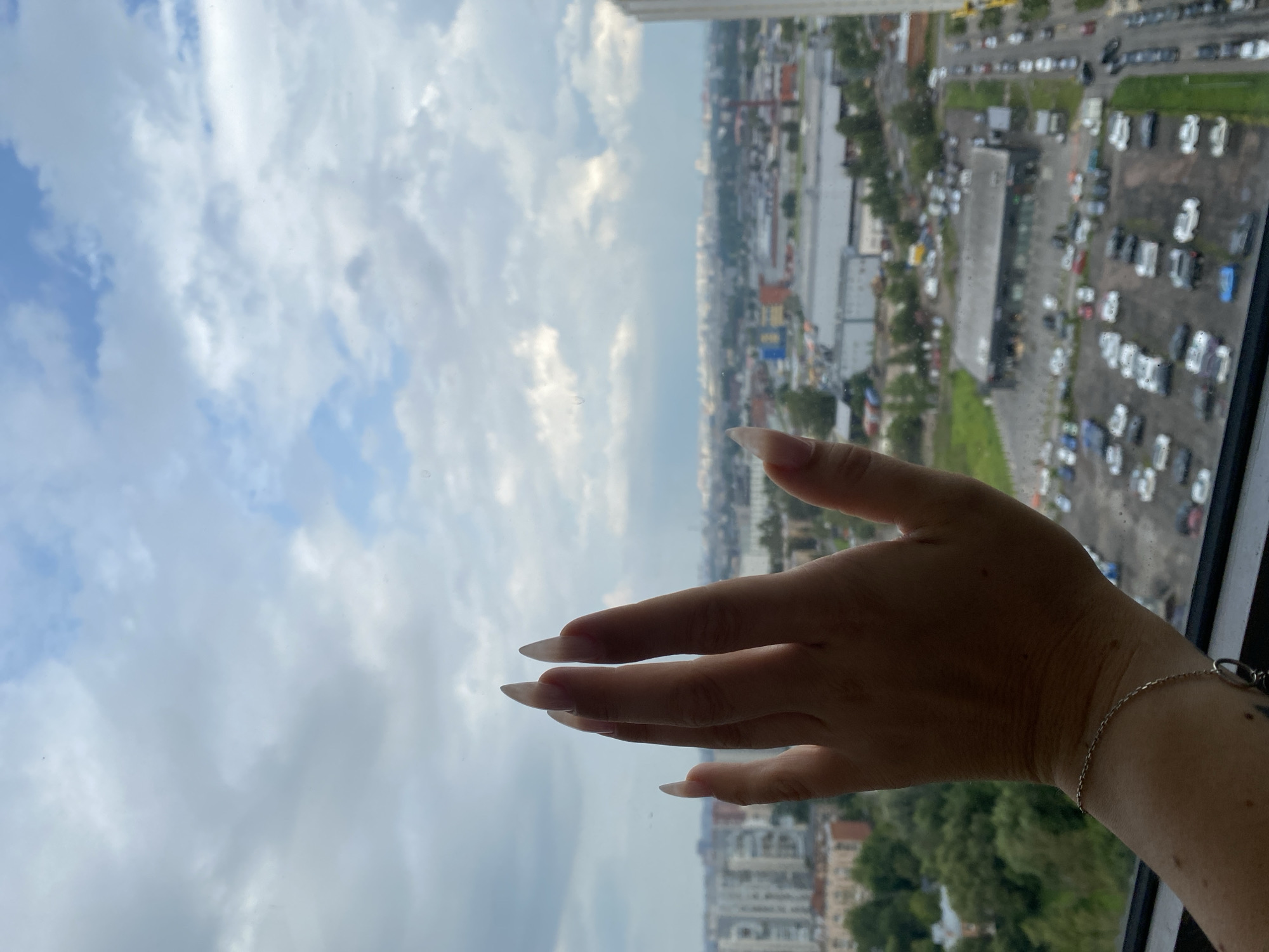 The image shows a close-up of a person's hand, with long, pointed fingernails, held up against a window. The hand is casting a shadow on the glass. Through the window, a cityscape is visible, including buildings, trees, and a parking lot filled with cars. The sky is cloudy and overcast. The location is identified as Ostrovki, Russia. The image appears to be taken from a high vantage point, possibly a high-rise building, looking out over the urban landscape. The overall tone is neutral, with no specific emotional cues.