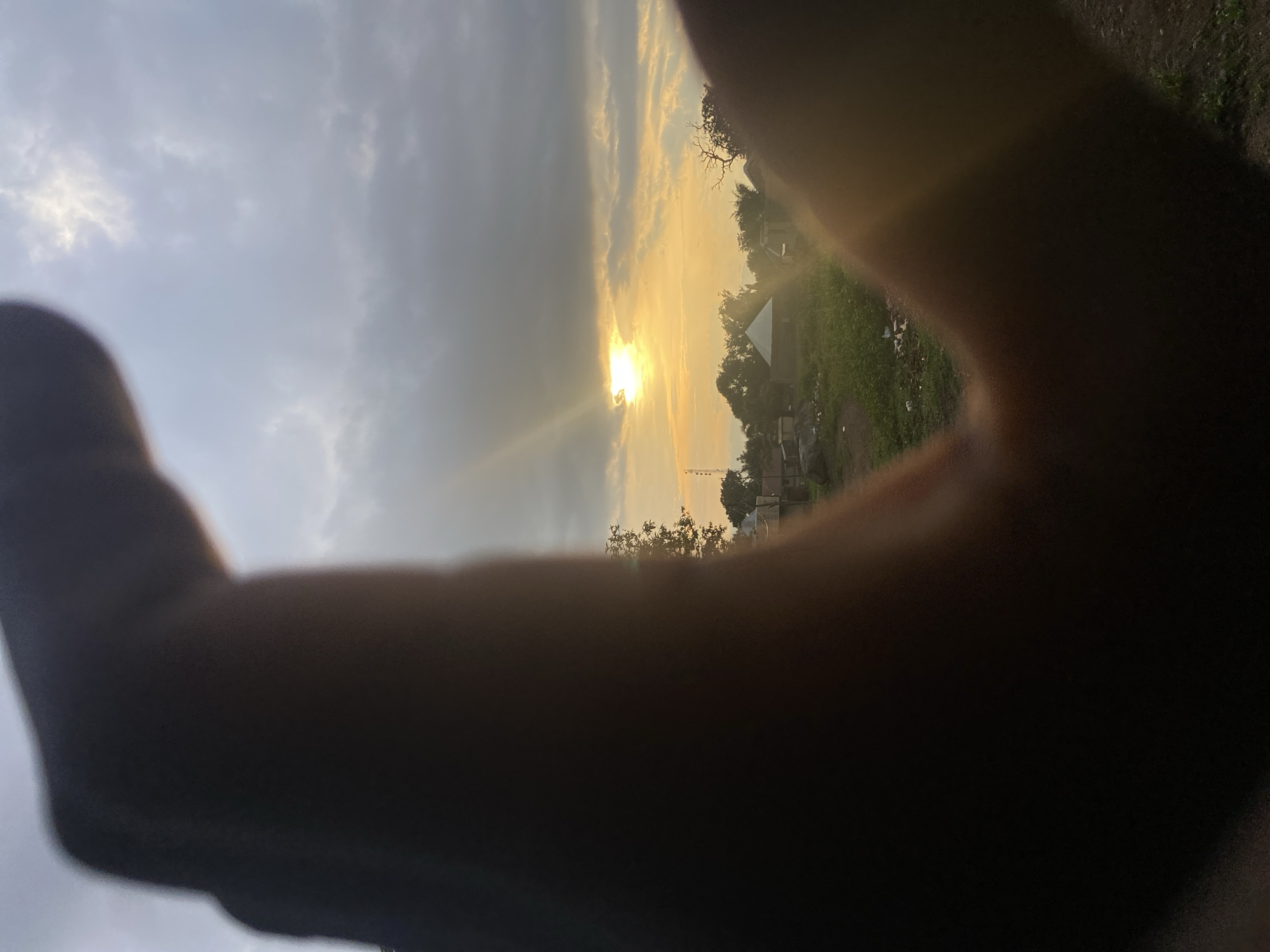 The image depicts a partially obscured view of a sunset in what is presumed to be Mararaba, Nigeria, though definitive landmarks are absent. The sun, partially obscured by clouds, is visible. A blurred foreground is dominated by what appears to be a human hand forming a framing gesture. Beyond the hands, a hazy landscape unfolds, revealing scattered buildings amidst greenery. A communications tower can be seen, The environmental lighting suggests either early morning or late evening.
