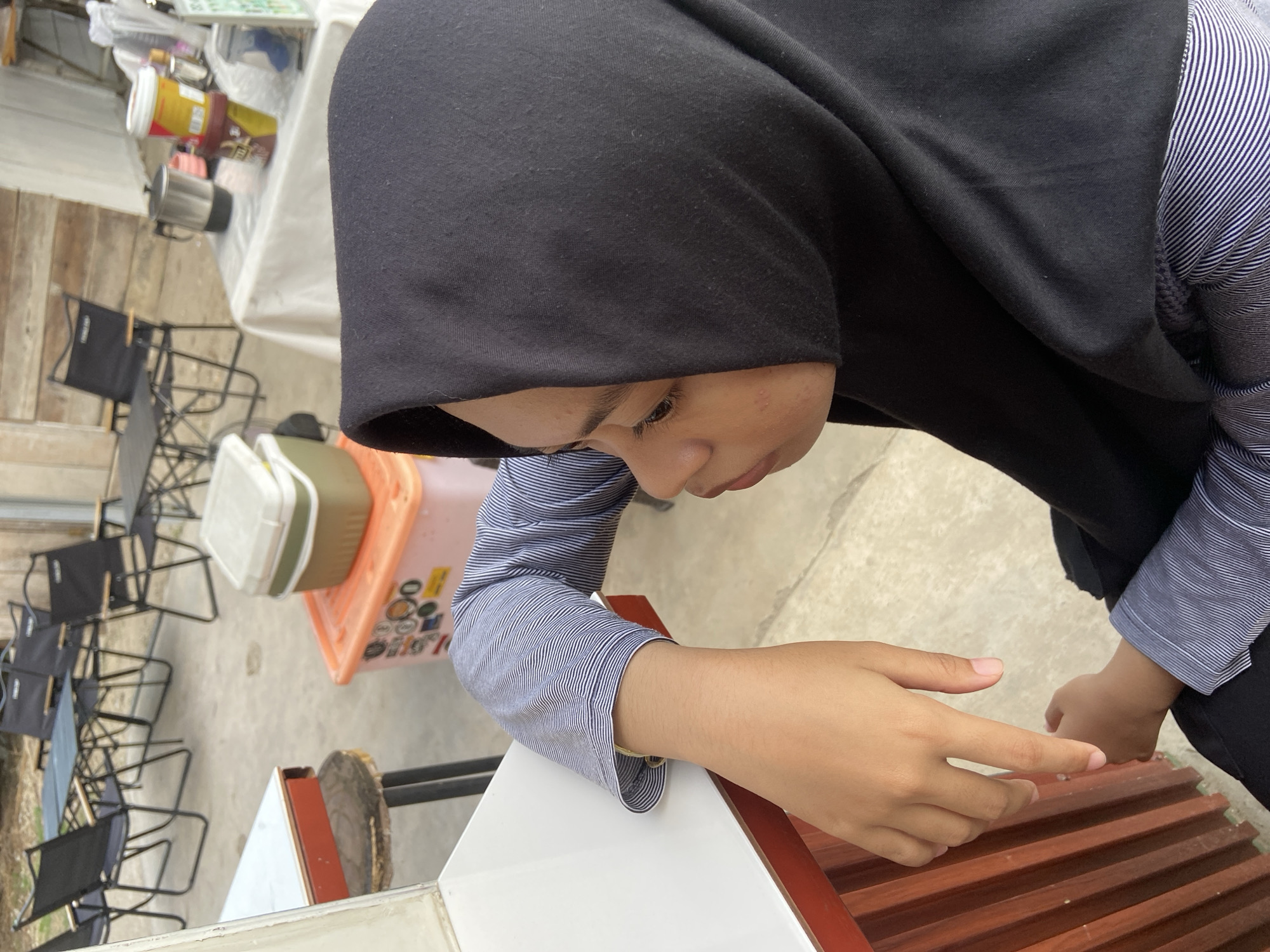 A young woman wearing a black hijab and a blue and white striped long-sleeved shirt is leaning on a white counter. Her left arm is resting on the counter, and her hand is dangling. Her right arm is bent, and her hand is resting on her lap. She is looking down and to her right. In the background, there are several folding tables and chairs set up on a concrete patio. There is also a cooler, an orange plastic bin with stickers, and a table covered with a white tablecloth with various items on it, including a coffee maker and jars. The scene appears to be an outdoor seating area, possibly a cafe or restaurant, in Tanjung Gading, Indonesia.