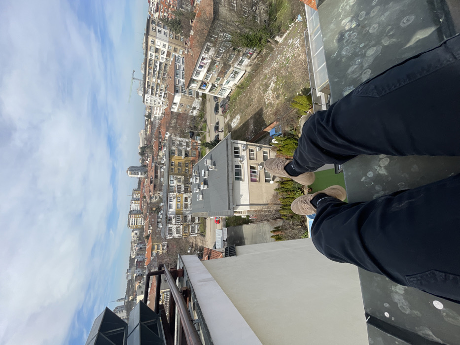 The image depicts a first-person perspective from an elevated position, likely a rooftop, overlooking a densely populated urban area. In the foreground, the lower half of a person's body is visible, wearing dark pants and light brown athletic shoes, with legs extended forward over the edge of a flat, grey, weathered surface. White circular marks are visible on this surface. To the immediate left, a white parapet wall is present, with industrial-style metal structures, possibly solar panels or ventilation equipment, partially visible further left.
The background presents a panoramic view of a city under a partly cloudy sky with patches of blue. The urban landscape consists primarily of multi-story residential buildings, varying in age and architectural style. Many structures feature red-tiled roofs and beige or white facades, interspersed with buildings having flat, grey roofs. Streets with parked and moving vehicles are visible winding through the urban fabric. Scattered trees, some bare and some with green foliage, line the streets and residential properties. In the middle-right distance, a tall construction crane indicates ongoing development. Further in the background, hazy outlines of hills or mountains are discernible. The scene is located in Sofia, Bulgaria.