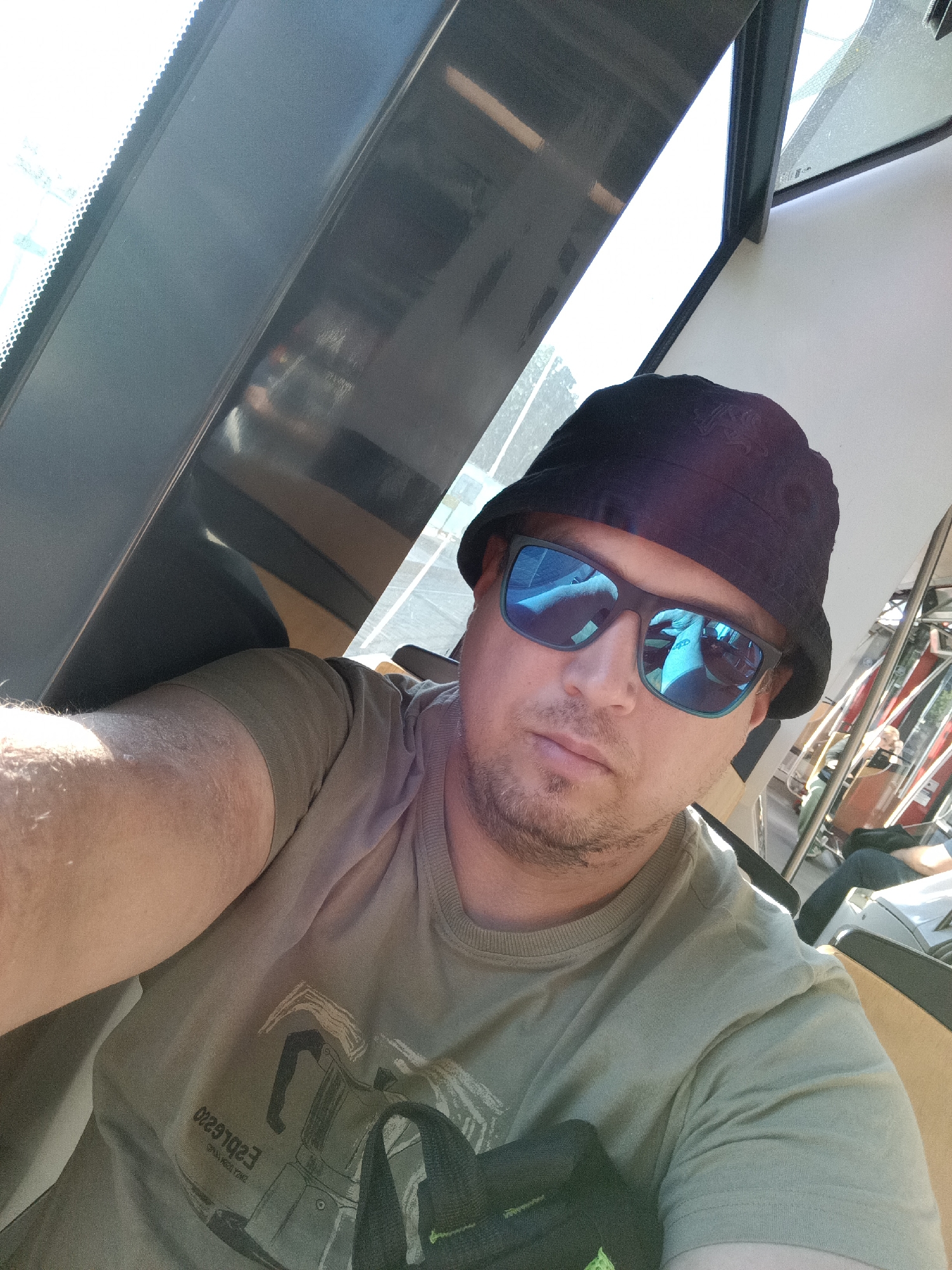 A man wearing sunglasses and a bucket hat is taking a selfie in what appears to be public transportation. The image is taken from a selfie perspective, with the man looking directly at the camera. He is wearing a green t-shirt with a graphic print and text. The background shows the interior of a vehicle, with seats and windows visible. The lighting suggests it is daytime. No specific landmarks of Prague are visible in the image.