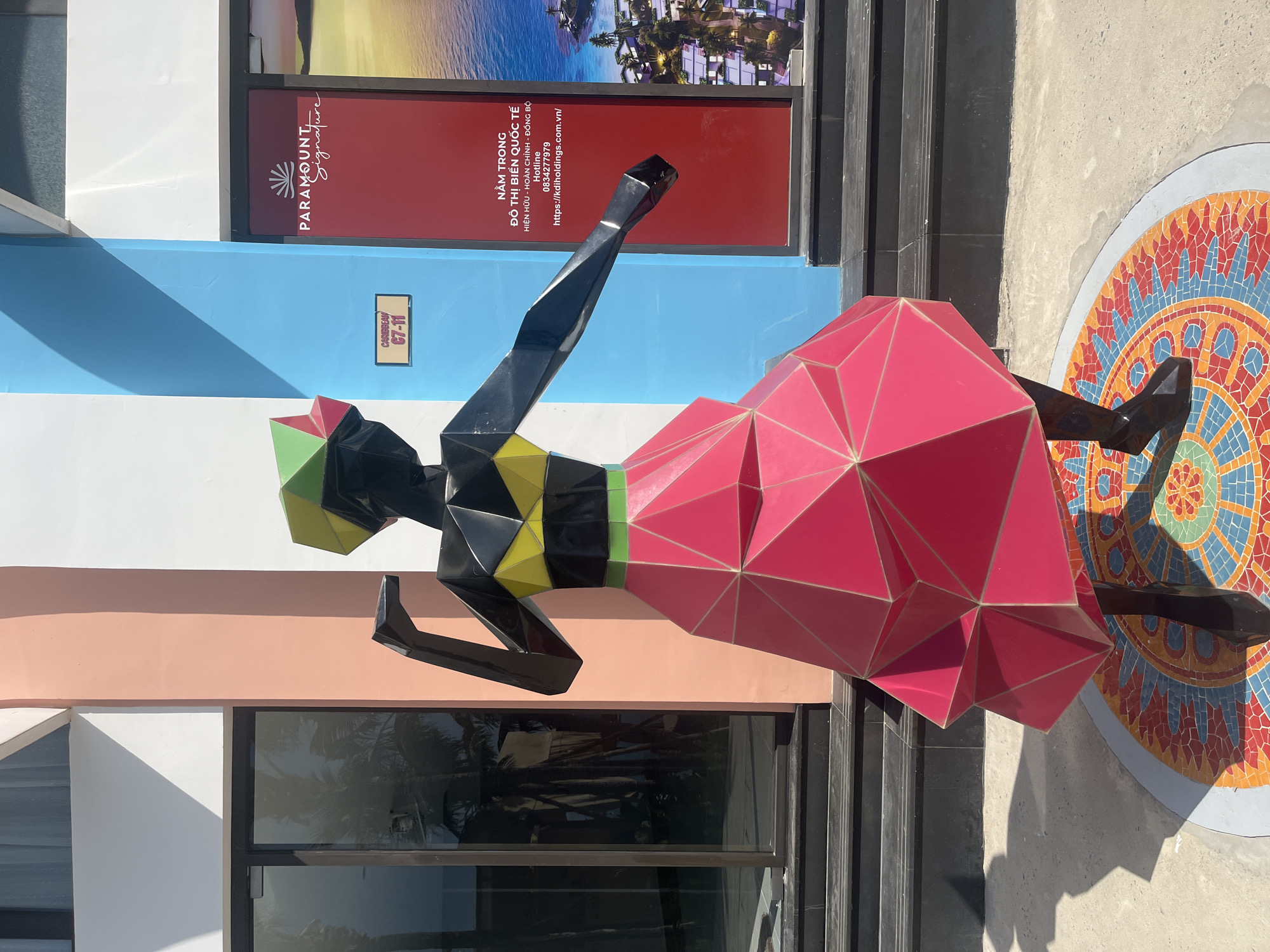 This image depicts a vibrant outdoor sculpture of a person dressed in a geometric-patterned outfit, standing on a circular mosaic base. The sculpture is black with a brightly colored triangular skirt, a yellow band around its chest, and a multi-colored geometric headpiece. The background features a modern building with large glass doors and walls painted in distinct pastel colors: peach, blue, and white. A red sign with white text, including the "PARAMOUNT Signature" logo and Vietnamese text, is visible on the building. The sculpture is positioned on a light-colored paved surface, with shadows cast by the sun. The mosaic base features a colorful mandala-like design with red, orange, yellow, blue, and green tiles. The environment suggests a public space, likely an entrance to a commercial establishment or resort. The style of the sculpture and the building suggests a contemporary artistic installation. Based on the Vietnamese text on the sign, which includes "ĐÔ THỊ BIỂN QUỐC TẾ" (International Coastal City) and a phone number often associated with Vietnamese businesses, and the context of "PARAMOUNT Signature," this location is likely within a resort or development in Nha Trang, Vietnam. The sign provides a hotline number (0834277979) and a website (https://kdiholdings.com.vn/), which are typical of real estate or hospitality projects in Vietnam.
