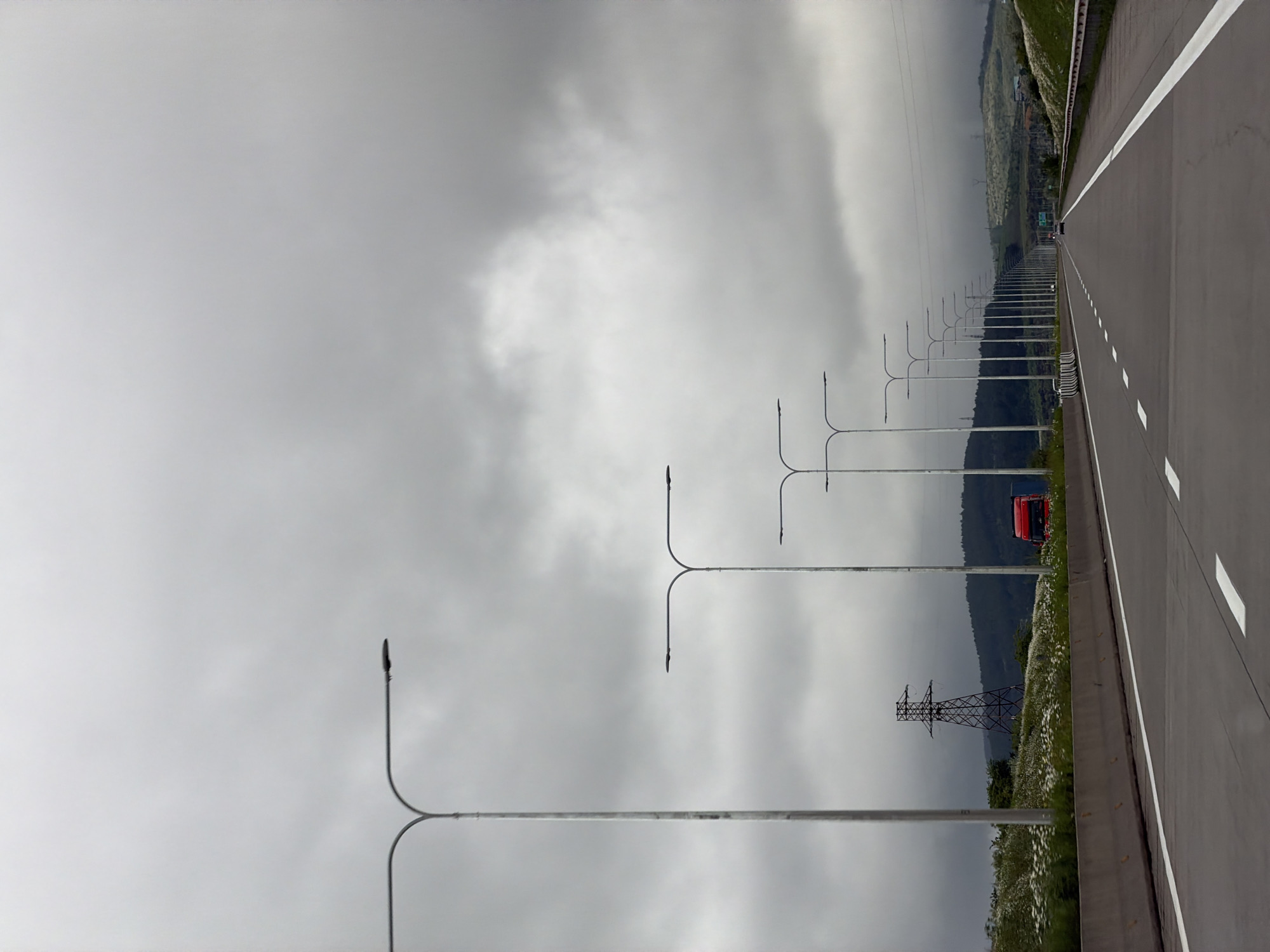 The image shows a long, straight highway under a cloudy sky in Ruisi, Georgia. The highway is marked with white lines and has a concrete barrier on the right. A row of evenly spaced streetlights runs along the side of the highway, disappearing into the distance. An electrical transmission tower is visible beside the highway. A red truck can be seen driving on the highway in the distance. The landscape beyond the highway barrier is characterized by grass and small green trees along the slope.