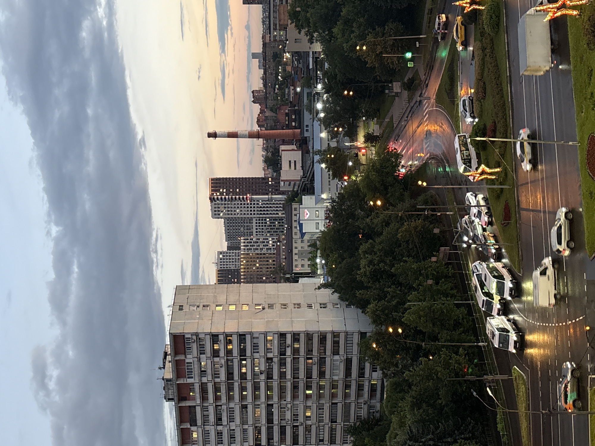 The image captures an urban scene in Pushkino, Russia, during what appears to be twilight or dawn. In the foreground, a multi-lane road is wet and reflects the lights of street lamps and passing vehicles. Several cars and vans are visible on the road, some of which are illuminated.
Dominating the left side of the frame is a tall, multi-story residential building with numerous windows, some of which are lit, suggesting occupancy. Beyond this building, a cluster of taller, more modern buildings with illuminated windows form part of the cityscape. A prominent industrial element in the mid-ground is a tall, red and white striped smokestack, characteristic of industrial areas.
The environment includes dense green trees lining parts of the road and surrounding some buildings. The sky above is filled with dramatic clouds, with hints of soft orange and pink hues suggesting the presence of the sun below the horizon. The overall atmosphere is one of a transitioning day in a built-up urban environment.