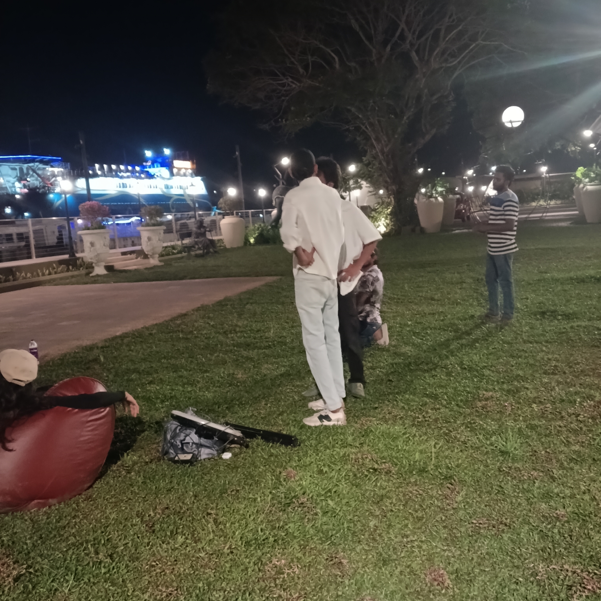 The scene captures a group of individuals in an outdoor setting at night, possibly in Panaji, India, based on the presence of a brightly lit cruise ship along the Mandovi River. A person wearing a hat and dark clothing reclines on a large, red beanbag chair, while a backpack and an object resembling a keyboard or lighting equipment lie on the grass nearby. Three other individuals stand together, two of them wearing white shirts and the third partially obscured behind them. A fourth person in a striped shirt and jeans stands further away. The location appears to be a park or garden area with manicured grass, decorative planters, trees, and illuminated streetlights.