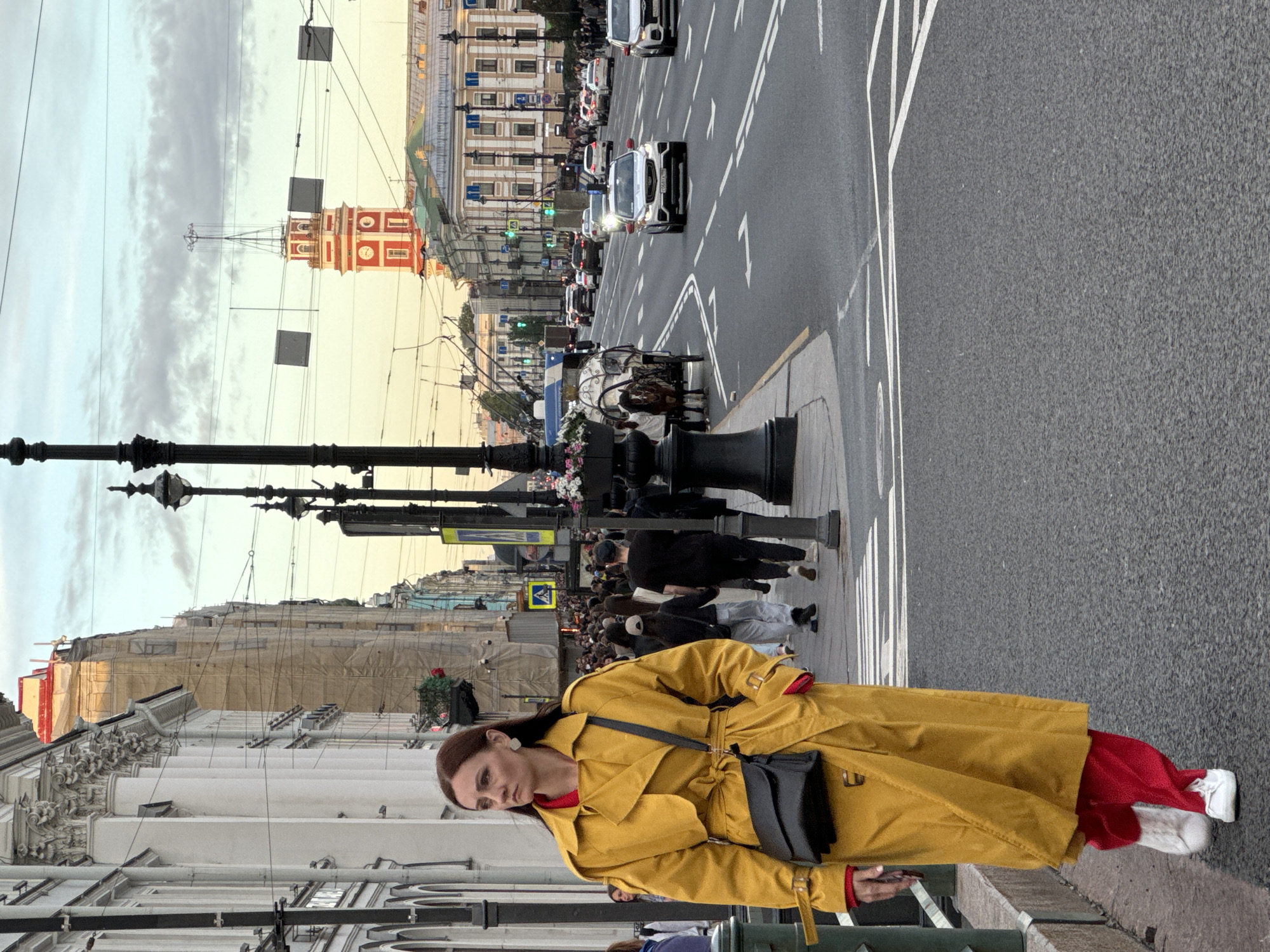 A medium shot captures a woman in a bright yellow trench coat and red pants walking on a crosswalk in Saint Petersburg, Russia. She carries a black purse slung across her body. Her gaze is directed to the left, away from the camera. In the background, a horse-drawn carriage is visible, along with a busy street filled with cars and pedestrians. Tall, ornate buildings line the street, and a prominent red clock tower stands in the distance. The sky is overcast with hints of sunset. The scene depicts a bustling urban environment with a blend of historical architecture and modern city life.