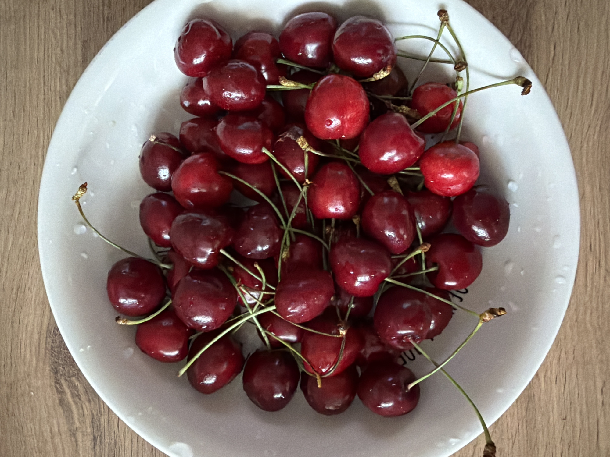 A close-up, top-down shot shows a white bowl filled with dark red cherries on a wooden surface. The bowl is white with a slightly textured rim and some water droplets on its inner surface. The cherries are plump, glossy, and have long, thin green stems, many of which are visible. Some cherries are clustered together, while others are spread out within the bowl. The lighting is natural, casting soft shadows and highlighting the wet surface of the cherries. The wooden surface beneath the bowl has a natural grain pattern and appears to be a table or countertop. No people are visible in the image. The scene is set in a kitchen or dining area in Chaikovsky, Russia.
