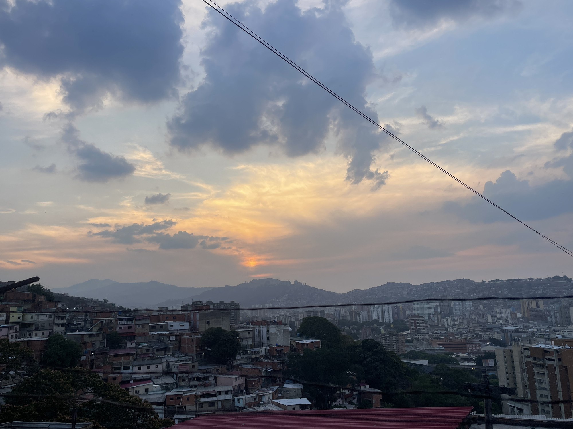 The image presents a high-angle view of a cityscape at dusk, most likely in Caracas, Venezuela. The foreground shows densely packed, low-rise buildings typical of informal settlements (ranchos), with a mix of brick, concrete, and corrugated metal roofs. The middle ground features a more established urban area with a combination of mid-rise apartments and commercial buildings. In the background, a series of hills are visible, partially obscured by haze or smog, with buildings extending onto their slopes. The sky is filled with a mix of dark, ominous clouds and lighter, orange-tinted clouds near the horizon, indicating the setting sun. Two parallel power lines stretch across the upper portion of the frame. There are no people visible in the frame.