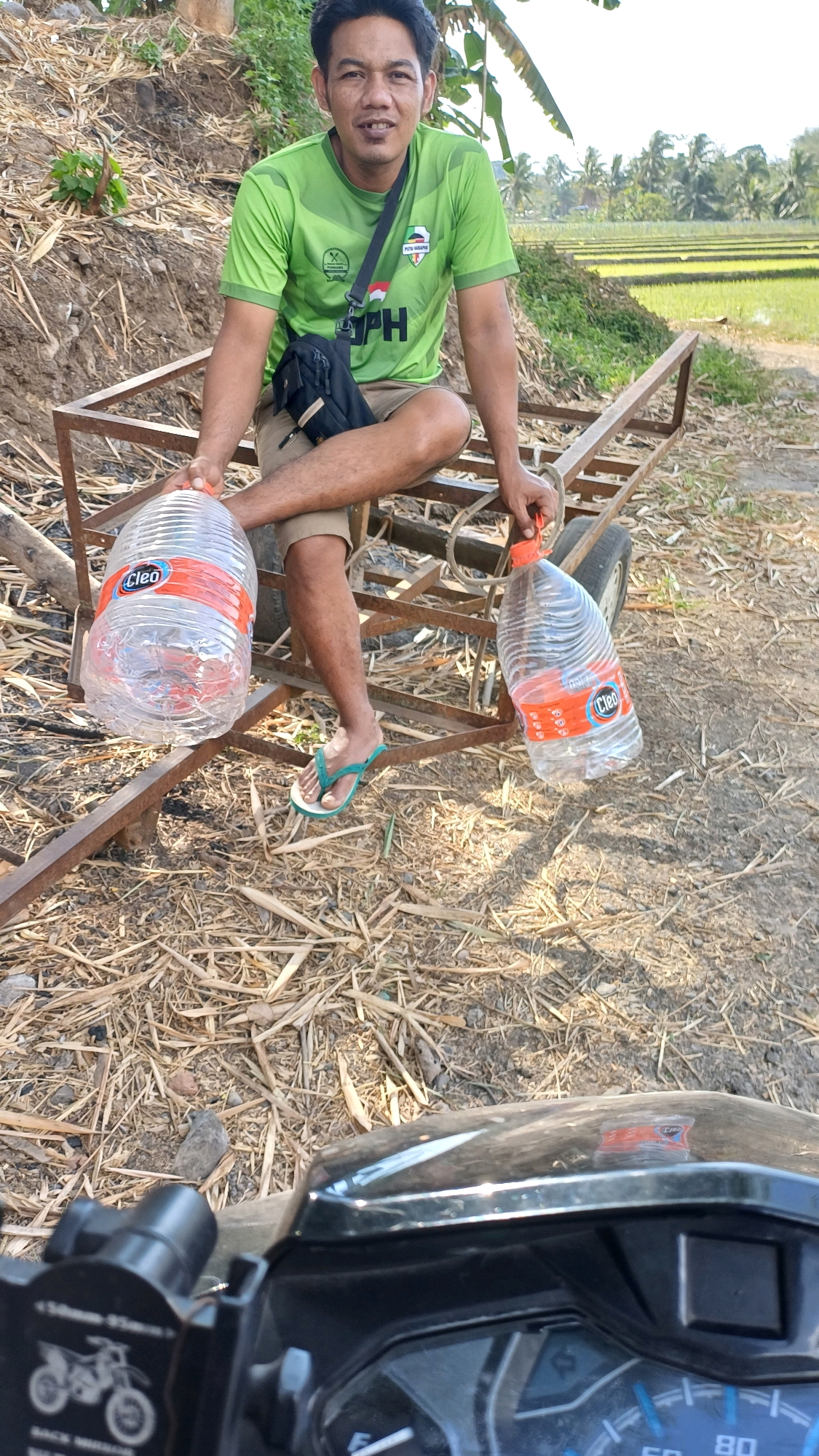 A man is sitting on a metal cart in an outdoor setting. He is wearing a green t-shirt, brown shorts, and flip-flops. He is holding two large plastic water bottles, one in each hand. The bottles have orange labels with the word "Cleo" written on them. The background shows a grassy field with some trees. The man appears to be smiling at the camera. The scene is likely in the rural area of Wonokerso, Indonesia, given the agricultural landscape. The cart appears to be used for transporting goods. The ground is covered with dry leaves and straw. A motorcycle is visible in the foreground, suggesting the man may have arrived on it.