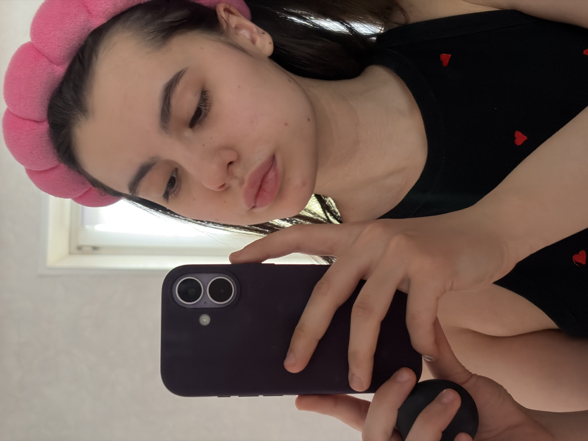 A young woman with a pink padded headband is taking a selfie with a purple-cased iPhone. She is wearing a black tank top with small red heart patterns. A window is visible in the background. The location, Chaikovsky, Russia, cannot be verified from the image.