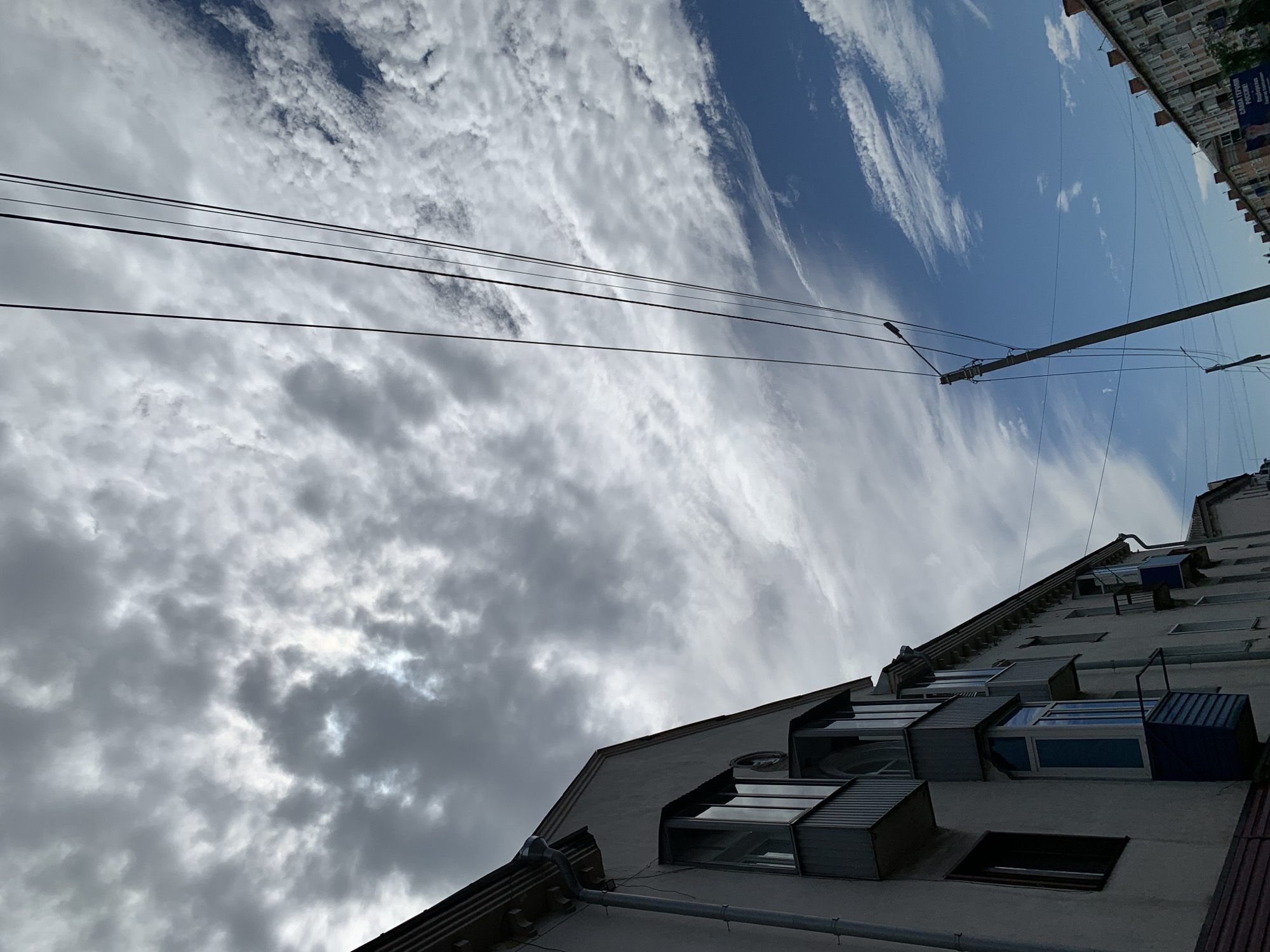 In Prokopyevsk, Russia, this image captures a street scene dominated by a dramatic sky. A light-colored apartment building with multiple balconies occupies the left side of the frame, its architecture partially obscured by the angle. Overhead, a dense collection of dark, textured clouds stretches across the upper portion of the image, interspersed with patches of brighter sky and a sliver of sunlight. Parallel to the building, several electrical wires extend diagonally upwards, cutting through the sky. A utility pole with a street light is visible on the right, leaning slightly, with more wires emanating from it. Further down the street on the right, another apartment building with a red-tiled roof and brick facade is visible, along with what appears to be a billboard advertising "СЛАВА ГЕРОЯМ РОССИИ!" (Glory to the Heroes of Russia!). The overall atmosphere is overcast with hints of blue sky peeking through, suggesting dynamic weather conditions.