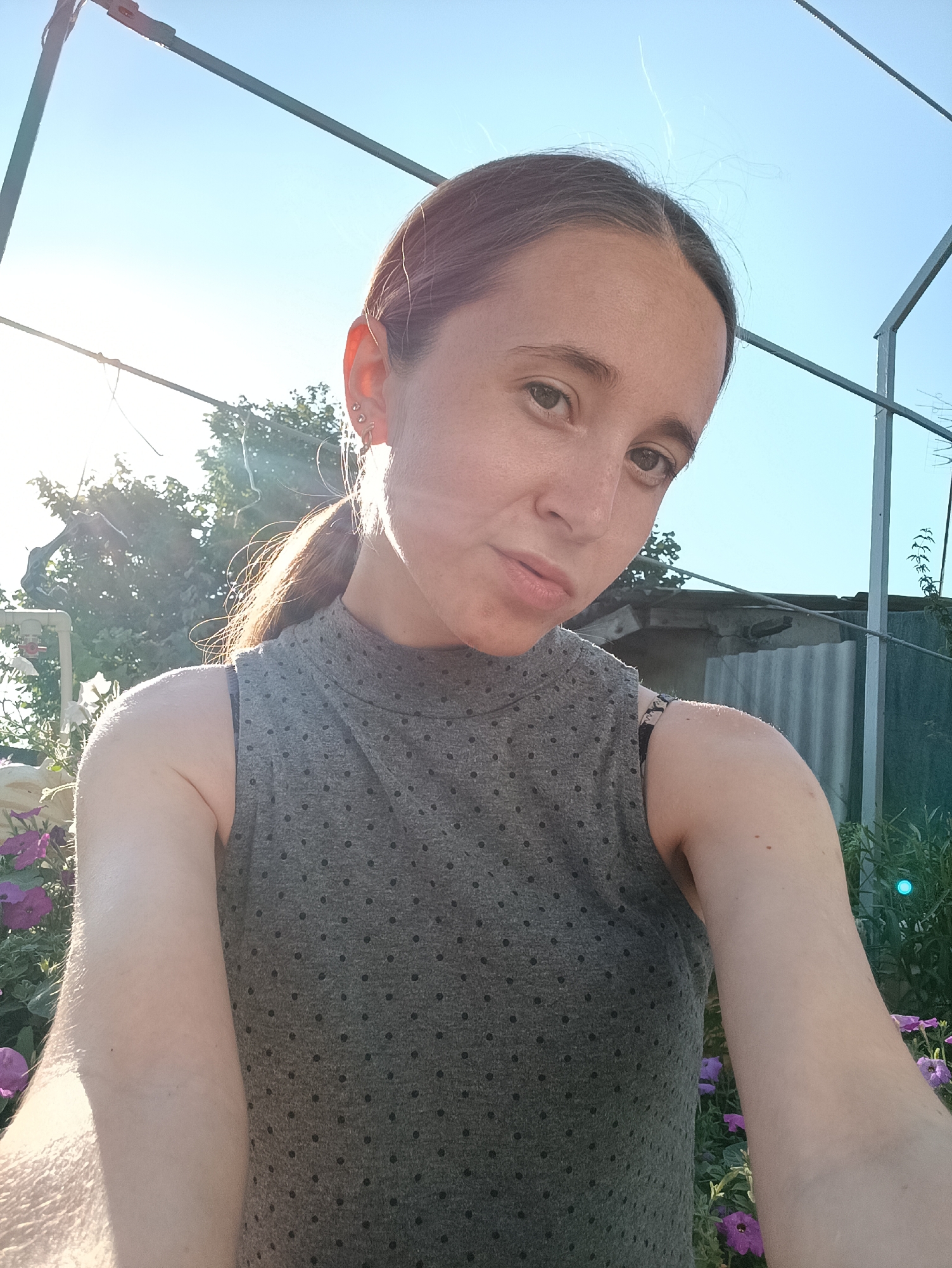 A medium shot selfie taken outdoors on a sunny day shows a young woman with her hair pulled back in a ponytail, looking at the camera. She is wearing a grey sleeveless top with a high neckline and a pattern of small black dots. The background includes a clear blue sky, some trees, and a corrugated metal fence. In the foreground, there are purple flowers. The image appears to have been taken in Vypasne, Ukraine, given the presence of what looks like a clothesline or some type of wire structure above the woman, which is common in rural Ukrainian backyards.