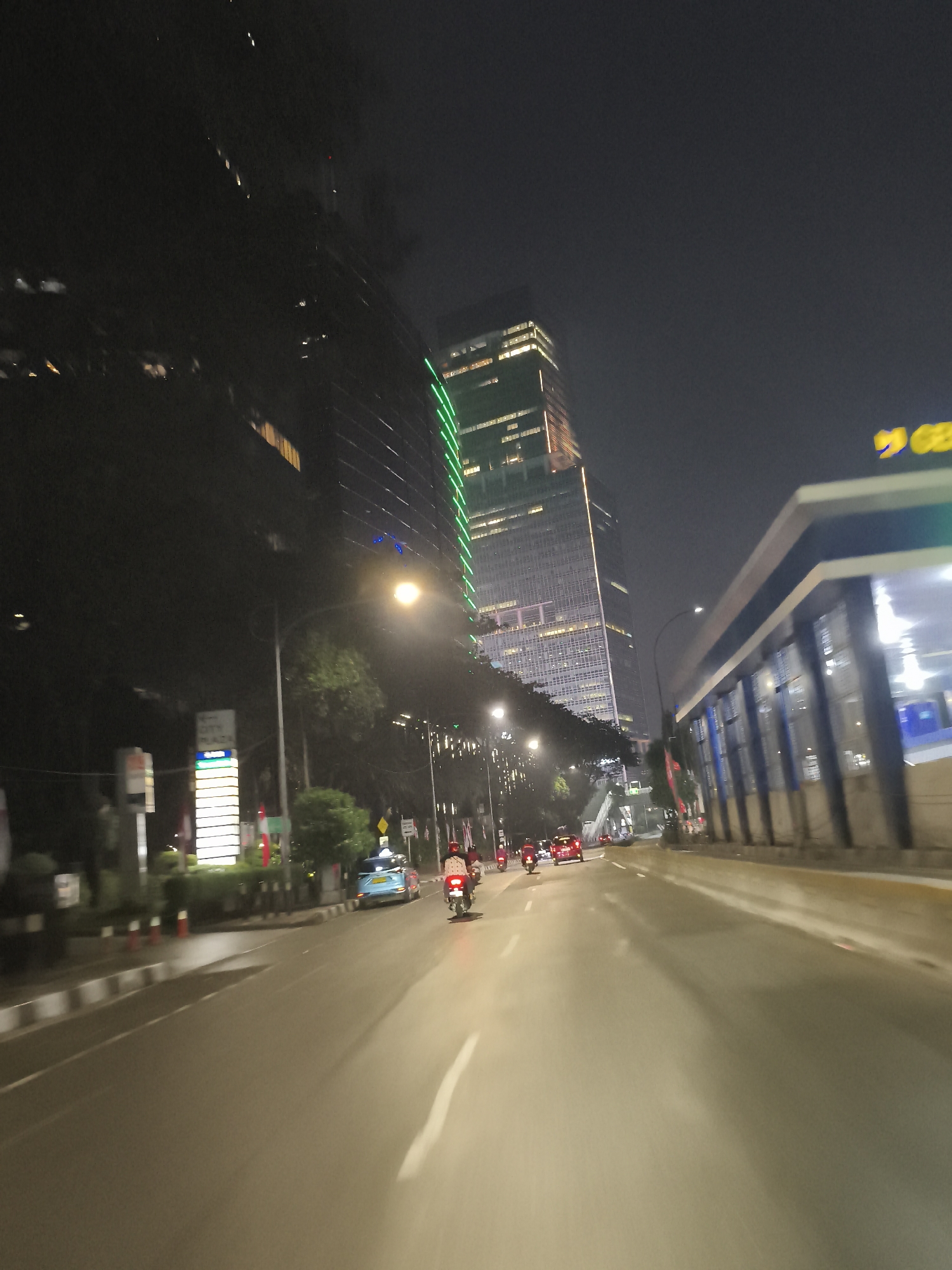 The image depicts a street scene at night in South Jakarta, Indonesia, possibly near Plaza Senayan. A tall, modern skyscraper with illuminated windows and vertical green lighting dominates the background. To the right, a building with a blue and white facade and an elevated walkway or structure is visible. The road in the foreground is wet, reflecting the streetlights and the headlights of vehicles. Several vehicles, including cars and motorcycles, are moving on the road. A blue car is in the middle lane, and motorcycles are on the left. A person is riding a motorcycle in the right lane. The overall atmosphere is that of a busy urban environment at night.
