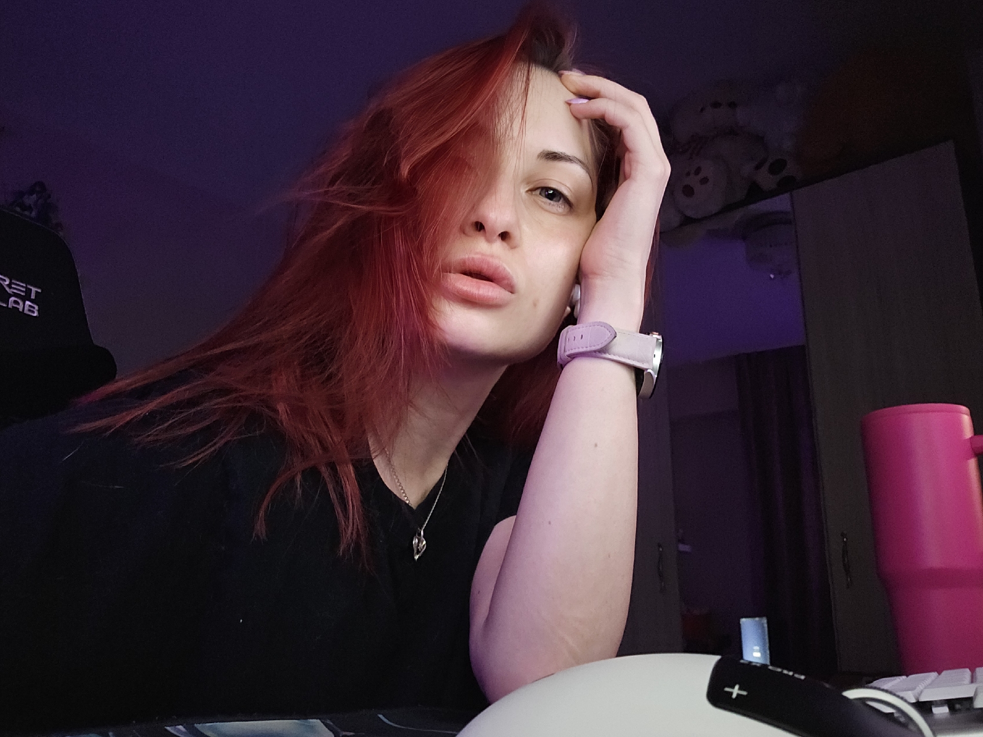 The image depicts an indoor scene in the city of Burgas, Bulgaria, characterized by dim lighting with a predominant purple ambient glow. A fair-skinned individual with long, reddish-orange hair is positioned centrally, gazing directly into the camera. A significant portion of her hair falls across the left side of her face, partially obscuring her left eye. Her visible right eye is dark, and her lips are slightly parted and appear full. Her left hand supports her head, with her elbow resting on a surface out of frame, forming a relaxed pose.
She wears a dark, possibly black, short-sleeved top, and a silver-colored necklace with a heart-shaped pendant is visible around her neck. On her left wrist, she wears a light lavender or pale pink wristwatch, and a white earbud is inserted into her left ear.
To the left of the individual, a portion of a black office chair is visible, bearing the white embroidered text "RET LAB" (suggesting "Secretlab"). In the background, positioned above and behind the individual's right shoulder, a shelf holds several light-colored plush toys, including at least one white teddy bear with visible paw print details. Adjacent to the plush toys, a dark-colored cabinet or piece of furniture is present. On the right side of the foreground, a vibrant pink insulated mug with a handle stands upright. Partially visible in the lower right, a white computer keyboard and a white and black computer mouse are positioned on a dark surface, likely a desk or mousepad.
