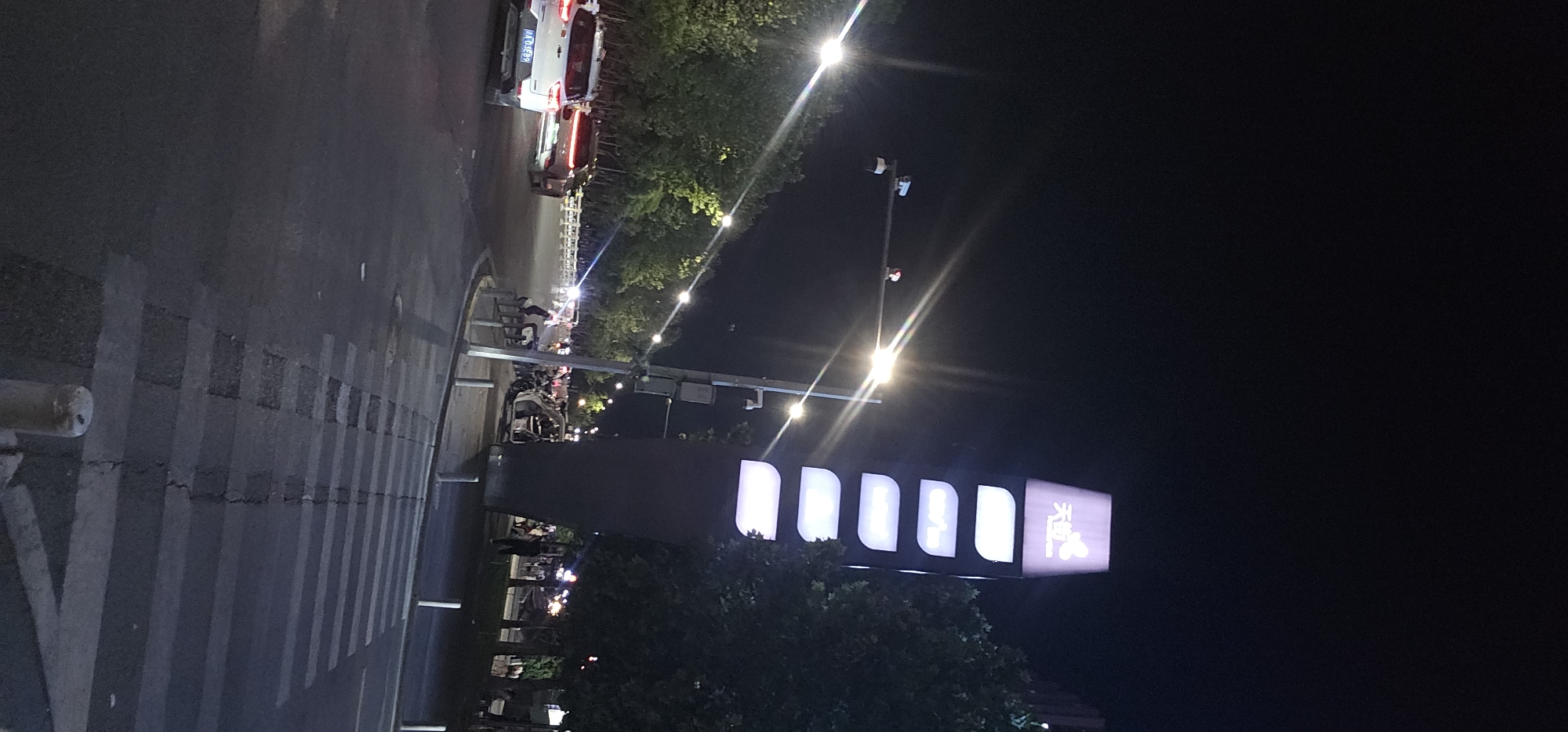 The image shows a street scene at night in the Chongqing High-tech Zone, China. A tall, illuminated signpost with multiple lit panels stands on the right side of the frame. In the foreground, a pedestrian crossing is visible on the asphalt road. Cars are driving on the street, including a white SUV with license plate A 03E89 on the left. Trees line the sides of the road, and streetlights cast illumination. Some people are present on the sidewalk and near the street, with activity suggesting a commercial or public area. The overall atmosphere is that of a busy urban environment at night.