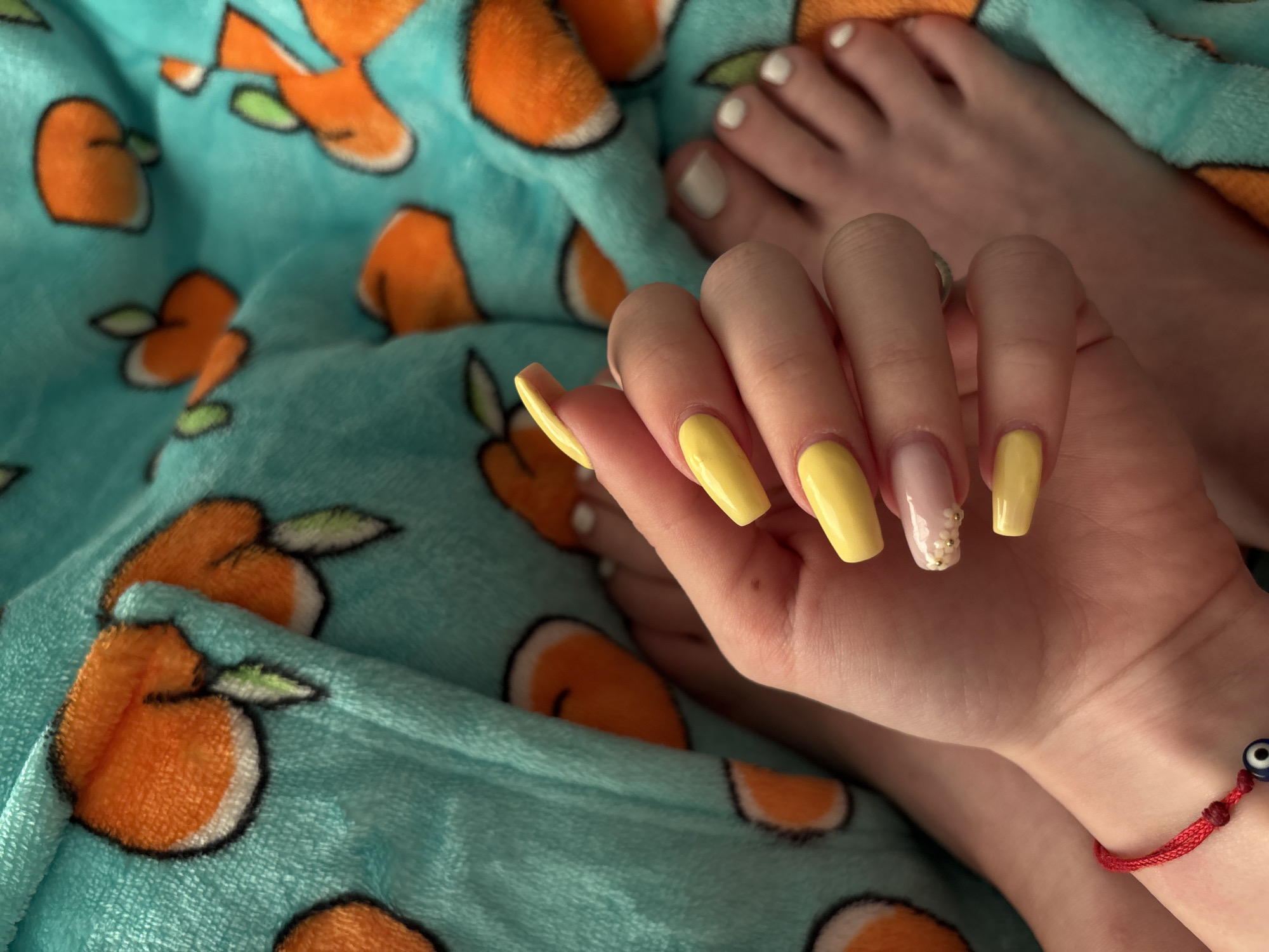 The image shows a person's hand with yellow manicured nails resting on a blue blanket with orange carrot patterns. The ring finger nail has a pink design with small white and gold embellishments. The person's toes are visible in the upper right corner, painted white. A red bracelet with a blue and white evil eye charm is visible on the person's wrist. The image does not provide any information about the location being Plovdiv, Bulgaria.