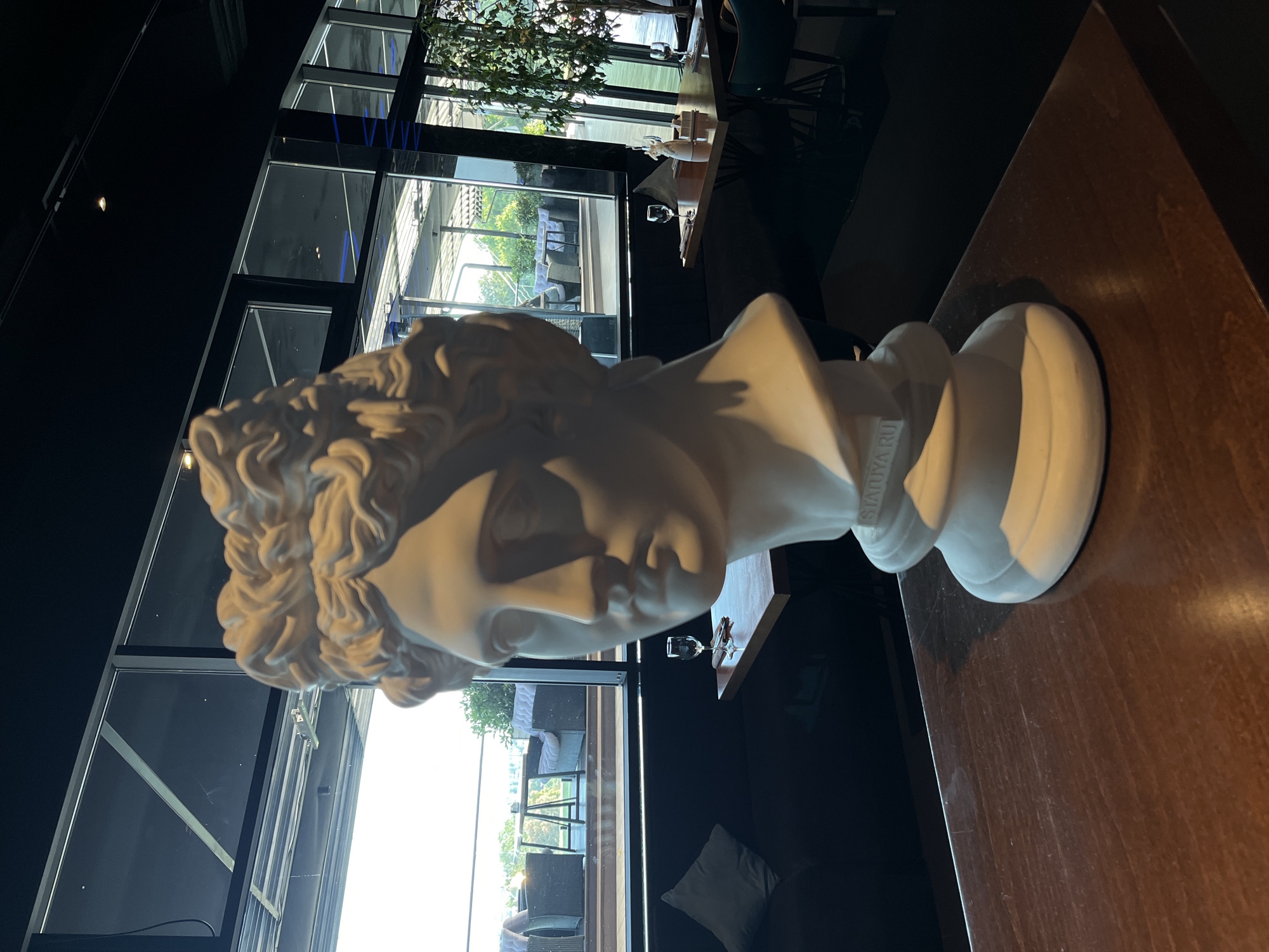 The image depicts a white plaster bust of a classical sculpture placed on a dark wooden table in a restaurant. The bust is positioned in the foreground, partially obscuring the view of the background. Behind the bust, through large windows, a view of an outdoor seating area with tables and chairs is visible, suggesting a restaurant or cafe setting. The windows also reflect the sky and trees, indicating it is daytime. The interior of the restaurant is dimly lit, with black seating and dark wooden tables. On one of the tables in the background, there are wine glasses and a napkin dispenser, further confirming the restaurant setting. The overall atmosphere appears to be a modern establishment with classical decorative elements. The location is identified as a restaurant in Sochi, Russia.
