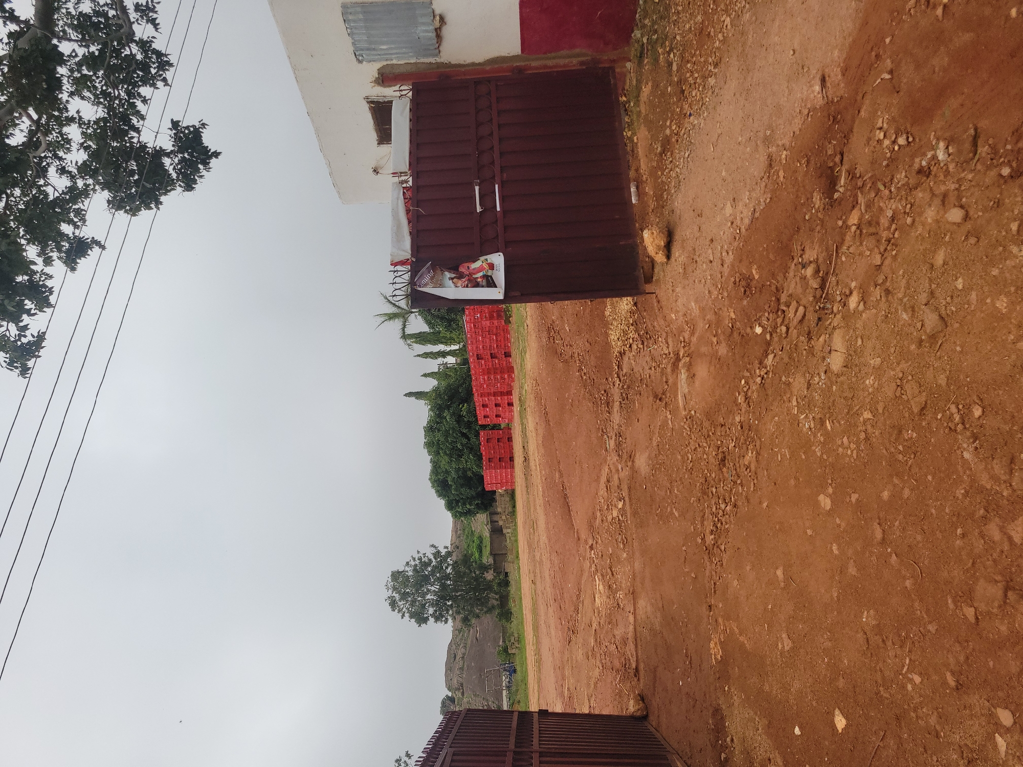 This image depicts the entrance to a facility in Jos, Nigeria. A large, dark red metal gate, partially open, is on the right side of the frame, with a poster attached to it. To the left of the gate is a white building with a red base, a window, and corrugated metal siding. Beyond the gate, a dirt and gravel road leads into an open area. In the background, a large pile of red plastic crates is visible, suggesting industrial or commercial activity. The terrain is uneven and dusty, with scattered rocks. In the far background, a rocky hill formation can be seen, along with trees and vegetation. The sky is overcast, with visible power lines overhead.