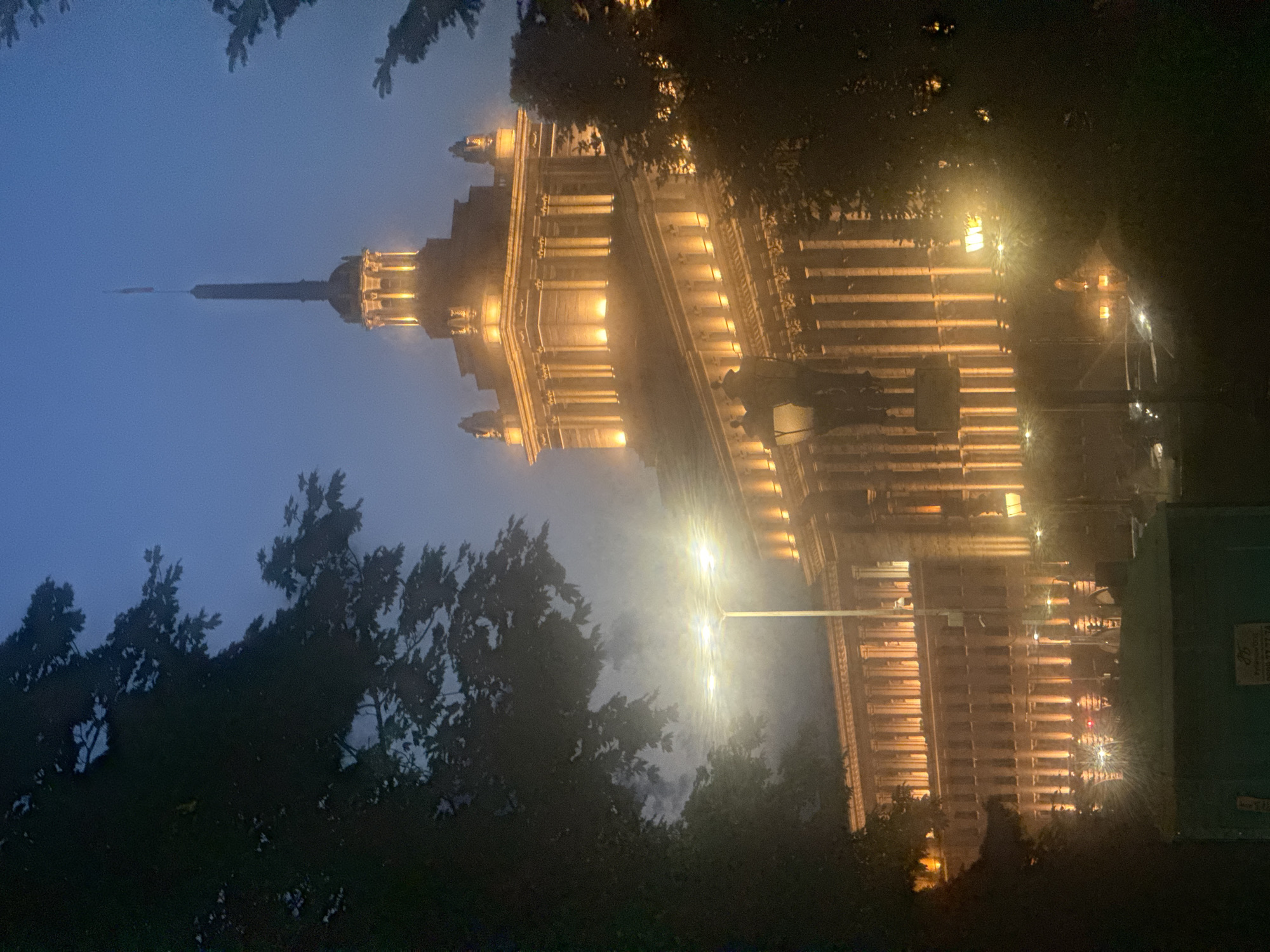 The image depicts the Largo, a central ensemble of administrative buildings in Sofia, Bulgaria, at dusk or night. The architecture is imposing, characterized by classical columns and illuminated windows that create a warm glow against the dusky blue sky. A prominent spire, topped with a flag, rises from one of the buildings. Trees frame the scene on either side, their dark foliage contrasting with the illuminated structures and a streetlamp that emits a bright, diffused light, suggesting the presence of fog or mist. The street in front of the buildings shows some car lights, indicating ongoing traffic. In the foreground, a dark green refuse bin is visible, with some text on its side. The overall atmosphere is somewhat atmospheric and perhaps a bit eerie due to the low light and fog.