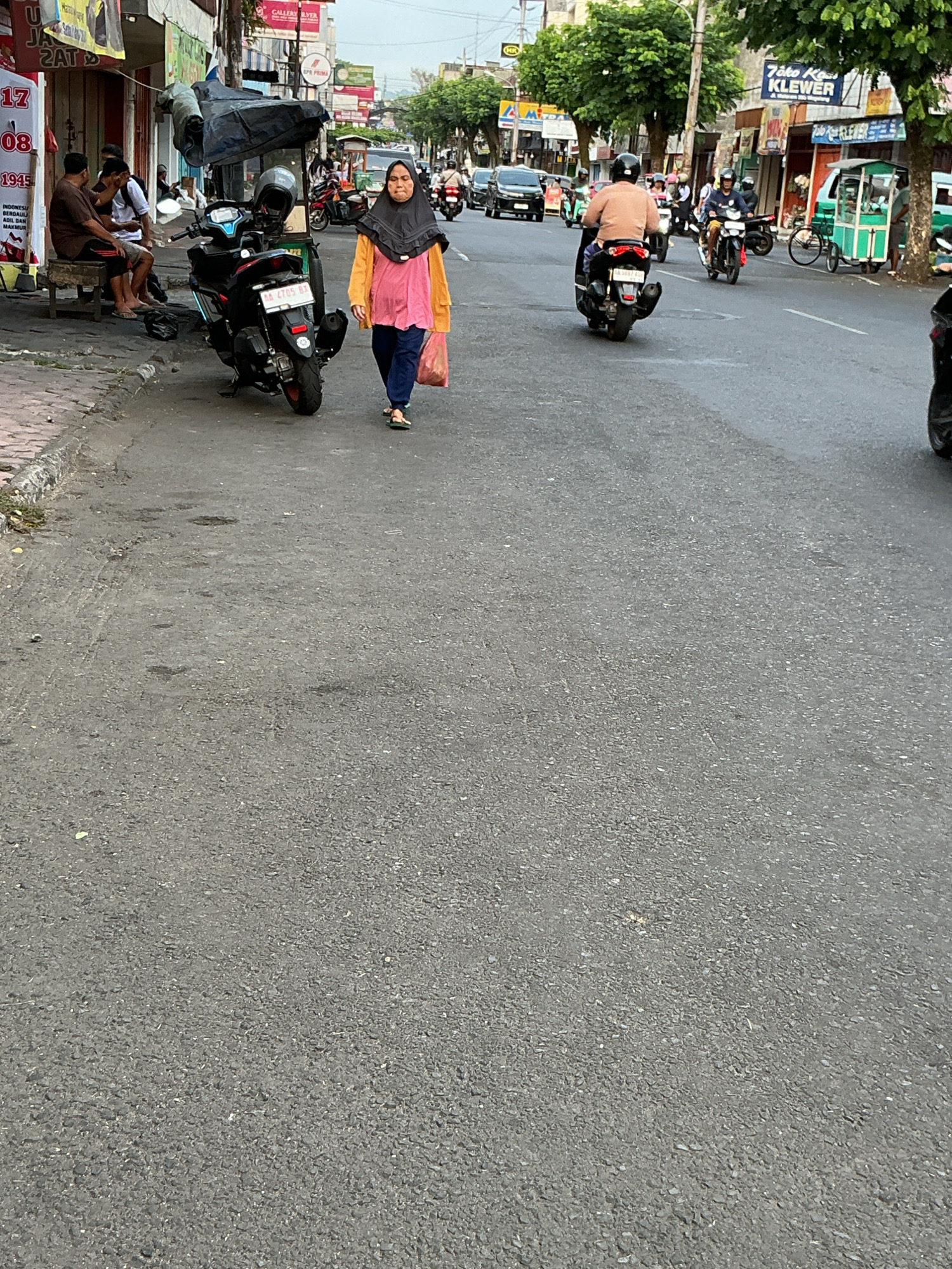 This is a street scene in Magelang, Indonesia. A woman wearing a hijab and an orange shirt walks on the asphalt road. She is carrying a pink plastic bag in her right hand. She is walking away from the viewer and towards the right side of the frame. Behind her, several motorcycles are parked along the side of the road, and a few people are sitting on a bench. Buildings line the left side of the street, displaying various signs and advertisements. In the background, more vehicles, including motorcycles and cars, are on the road. Trees and more buildings are visible further down the street. The sky is overcast.