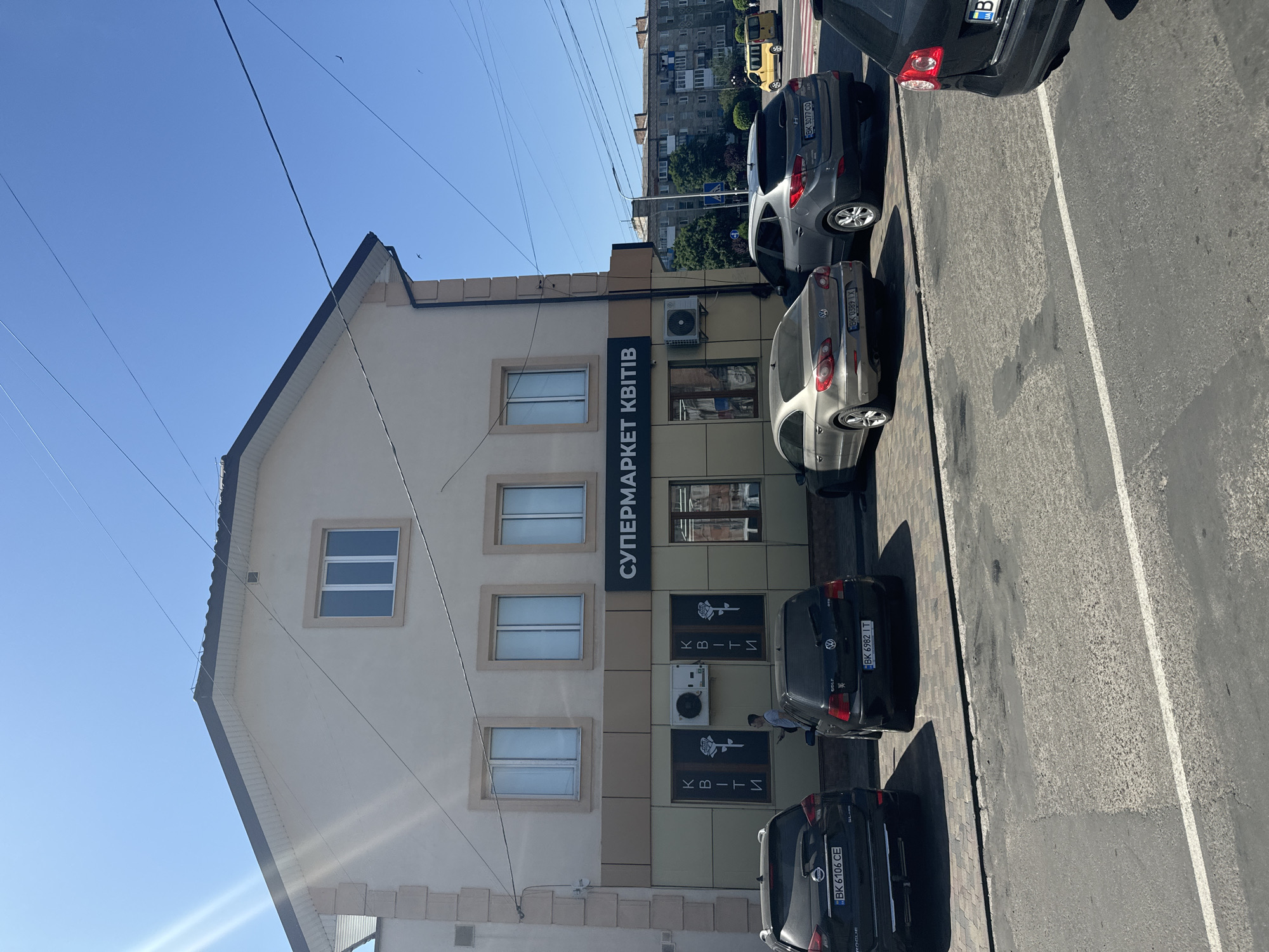The image shows a storefront in Sarny, Ukraine, labeled "СУПЕРМАРКЕТ КВІТІВ" (Flower Supermarket). The building is light beige with a dark grey roofline and tan-colored trim around the windows and lower facade. There are multiple windows on the upper and lower floors. The lower facade features two large windows, each with vertical text that reads "КВІТИ" (Flowers) and a stylized rose graphic. An outdoor air conditioning unit is mounted between these windows.
Several cars are parked in front of the building. On the left, a black Nissan Rogue with license plate BK 6106 CE is visible. Next to it is a black Volkswagen Golf with license plate BK 6982 IT. Further to the right, a silver Volkswagen CC with license plate BK 0389 IX is parked, followed by a grey Hyundai Tucson with license plate BK 3077 CO. A person in a light-colored shirt is standing near the air conditioning unit, holding a smartphone. In the background, apartment buildings and a yellow taxi are visible, suggesting an urban or suburban setting. The sky is clear and blue.