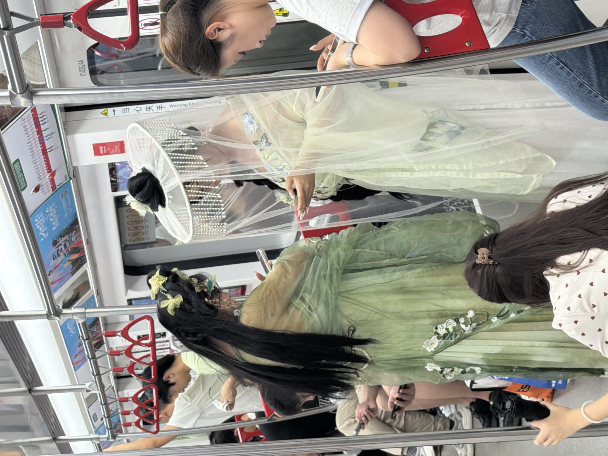 Inside a subway car, likely in Yuzhong District, China, two women are dressed in traditional Chinese Hanfu clothing. One woman, on the left, wears a pale green layered dress with floral embroidery at the hem and a long black wig adorned with yellow flowers and ornaments. She is holding a smartphone. The second woman, to her right, wears a white dress with a sheer, pale yellow overlay and an elaborate headdress adorned with beads and a veil. She is also holding a smartphone. Both women are the central focus, positioned amidst other subway passengers. A woman in a white t-shirt and blue jeans is visible to the right, also looking at her phone. The environment includes typical subway car features: metal poles, red grab handles, advertising posters along the ceiling, and doors. The overall scene captures a moment of cultural expression or perhaps a themed event within the public transportation setting.