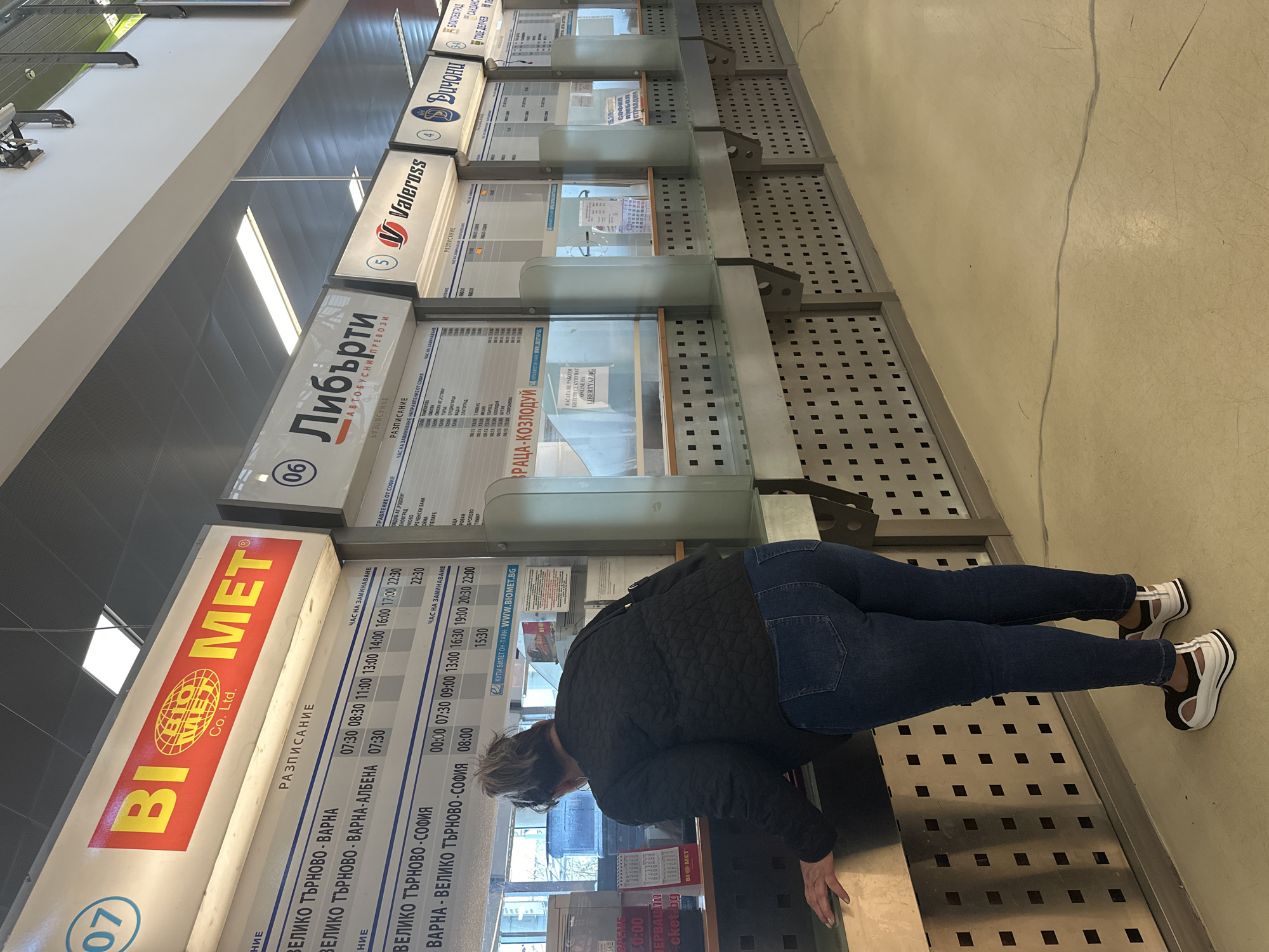 A person is standing at a schedule board in what appears to be a bus station or transit hub. The board, labeled "BIOMET Co. Ltd.", displays various departure times and routes, including destinations like Varna and Veliko Tarnovo. The schedules are in Bulgarian. Other schedules on the wall show other bus companies. The number "07" is in the bottom left corner. The location is Sofia, Bulgaria, based on the presence of Bulgarian text and the bus routes listed.