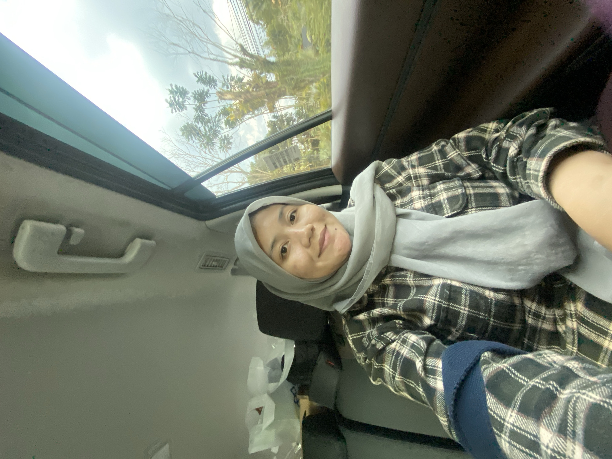 The image shows a woman wearing a grey hijab and a plaid shirt, sitting in the back seat of a vehicle. The interior of the vehicle is visible, including the car ceiling, seats, and a window. Outside the window, trees and power lines are visible, suggesting a setting in a populated area. The woman is looking towards the camera and smiling. Based on the visual cues, such as the vegetation and architectural style of nearby structures, the image was likely taken in Banjarbaru, Indonesia.