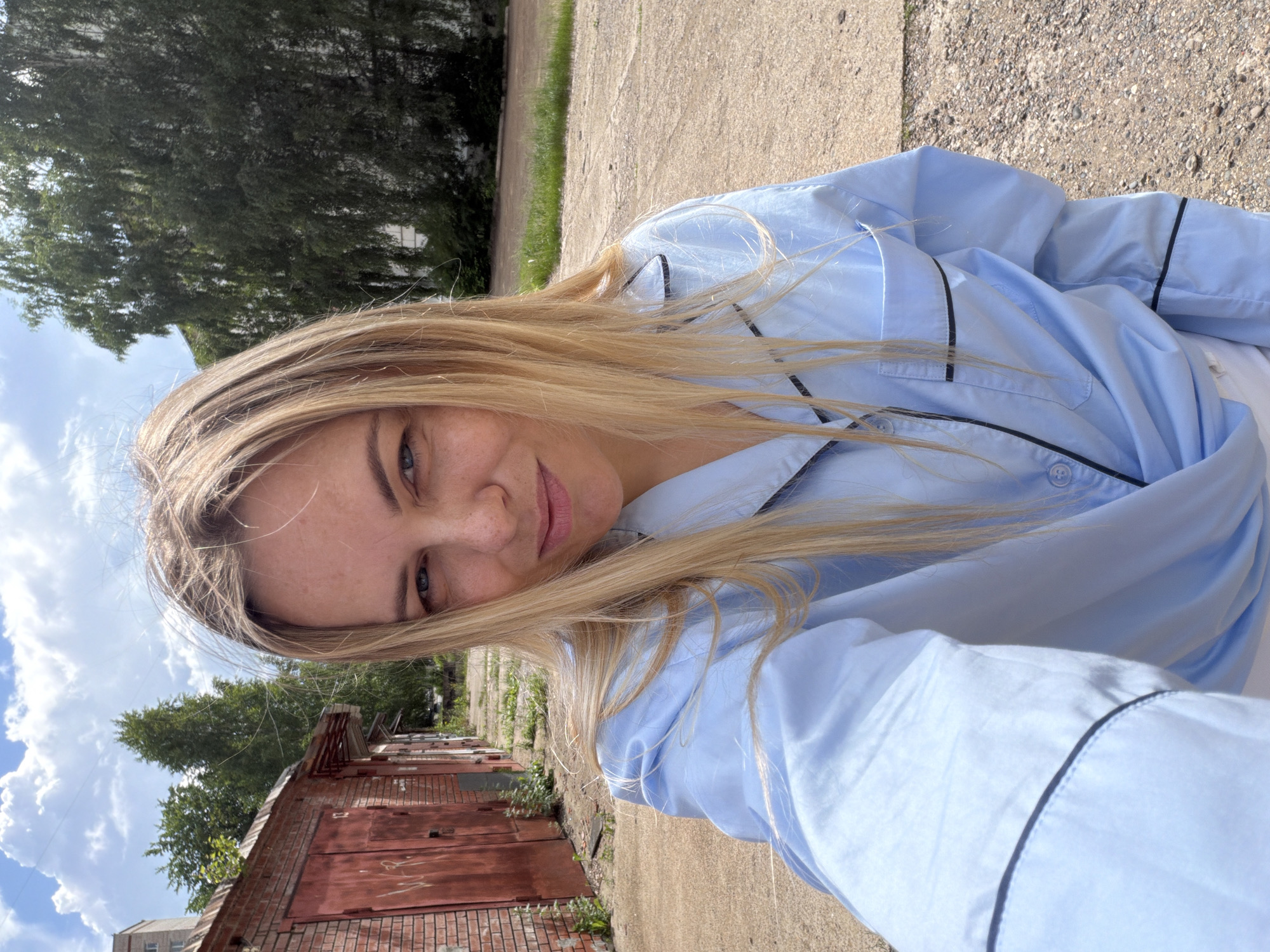 In Votkinsk, Russia, a woman with blonde hair and blue eyes takes a selfie outdoors. She is wearing a light blue pajama top with black trim. The background consists of a brick building with red garage doors and trees. The ground is a gravelly surface. The sky is partly cloudy.