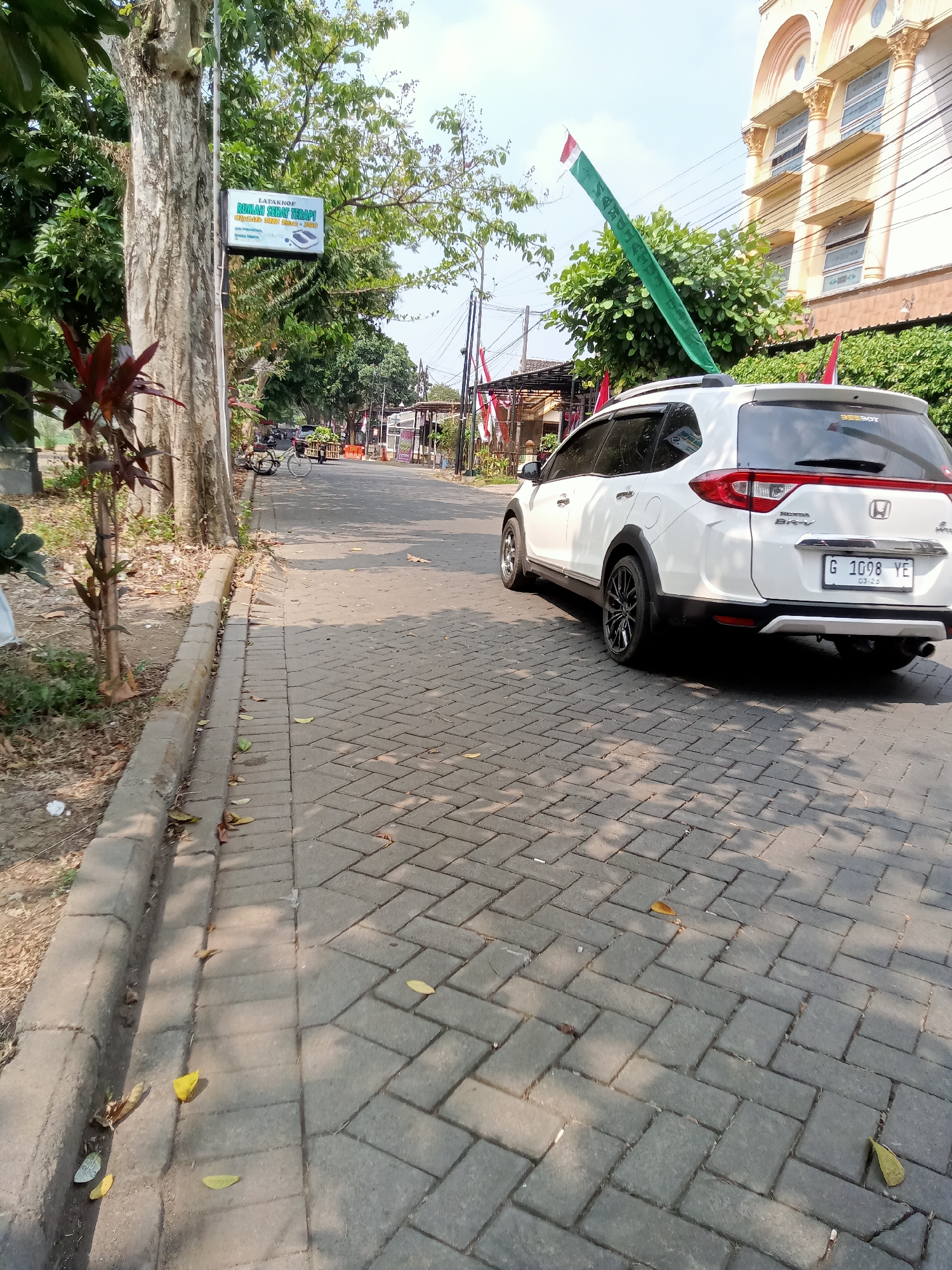 The image depicts a street scene in Moyoketen, Indonesia, on a sunny day. A white Honda BR-V is parked on the right side of the paved road, which is constructed with interlocking bricks. The car has a license plate that reads "G 1098 YE". A green flag with white text is attached to the roof of the car and extends upwards. To the left of the road, a sidewalk with a concrete curb is visible, lined with trees and some dry leaves on the ground. A sign with green text is mounted on a pole near one of the trees, and it appears to be advertising a health therapy service. Further down the street, there are buildings and other vehicles, including a bicycle. The environment is generally urban with some greenery.
