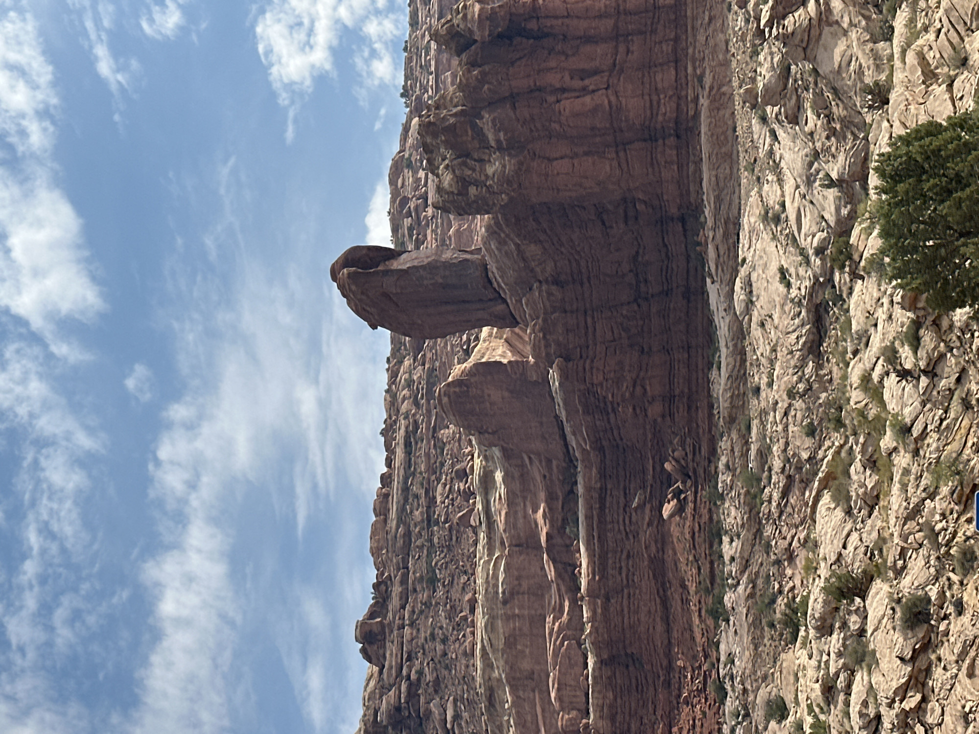 A vertical, medium-shot photograph captures a dramatic natural rock formation against a bright blue sky with scattered white clouds. The scene depicts a massive, layered rock cliff face, likely sandstone, exhibiting varying shades of brown and tan. The prominent feature is a large, mushroom-shaped rock that juts out horizontally from the cliff, appearing precariously balanced. Below this, another smaller rock formation juts out. The texture of the rock is rough and striated, suggesting erosion over time.
The lighting indicates it is daytime, likely late morning or early afternoon, with the sun casting shadows that highlight the contours of the rock. The weather appears clear and sunny. Vegetation is sparse, with small green bushes or trees clinging to the rocky surfaces in places. The scale of the rock formations suggests a grand, natural landscape, consistent with a desert or canyon environment. While no people are visible, the presence of a small blue sign on the far right edge suggests a possibility of a trail or viewing area, hinting at a tourist destination. The overall impression is one of natural wonder and geological spectacle. The location is identified as Moab, United States, a region known for its distinctive red rock formations and national parks.