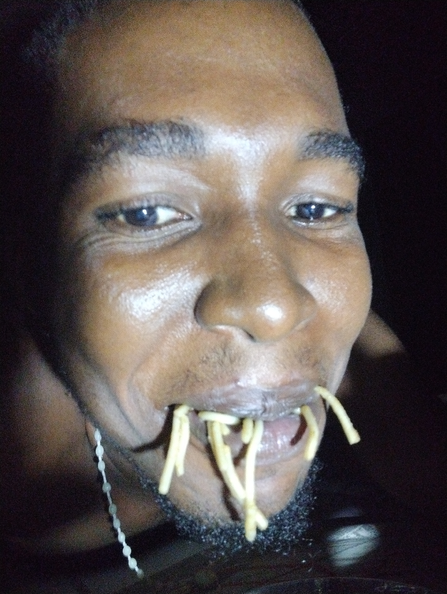This close-up portrait captures a dark-skinned individual, likely male, against a dark, undifferentiated background in Ihiagwa, Nigeria. The subject's face is illuminated by a direct flash, creating prominent highlights on the forehead, nose, and cheeks, suggesting low ambient light conditions, possibly at night or in a dimly lit indoor space.
The person is shown from above the forehead down to the neck. They have dark eyes, full dark eyebrows, and short, dark hair. A short beard or stubble frames the chin and mouth area. The most distinctive activity is the person holding or eating spaghetti or a similar type of pasta, with several pale yellow strands hanging out of their open mouth. Their expression appears playful or amused, indicated by a slight smile and a direct gaze towards the camera.
A thin, silver-colored chain necklace, featuring small, bead-like segments, is visible draped around the left side of the subject's neck. The skin has a somewhat shiny appearance, accentuated by the flash. The overall image quality suggests a candid or informal photograph taken with a flash, resulting in a slightly grainy texture. There is no visible text in the image.