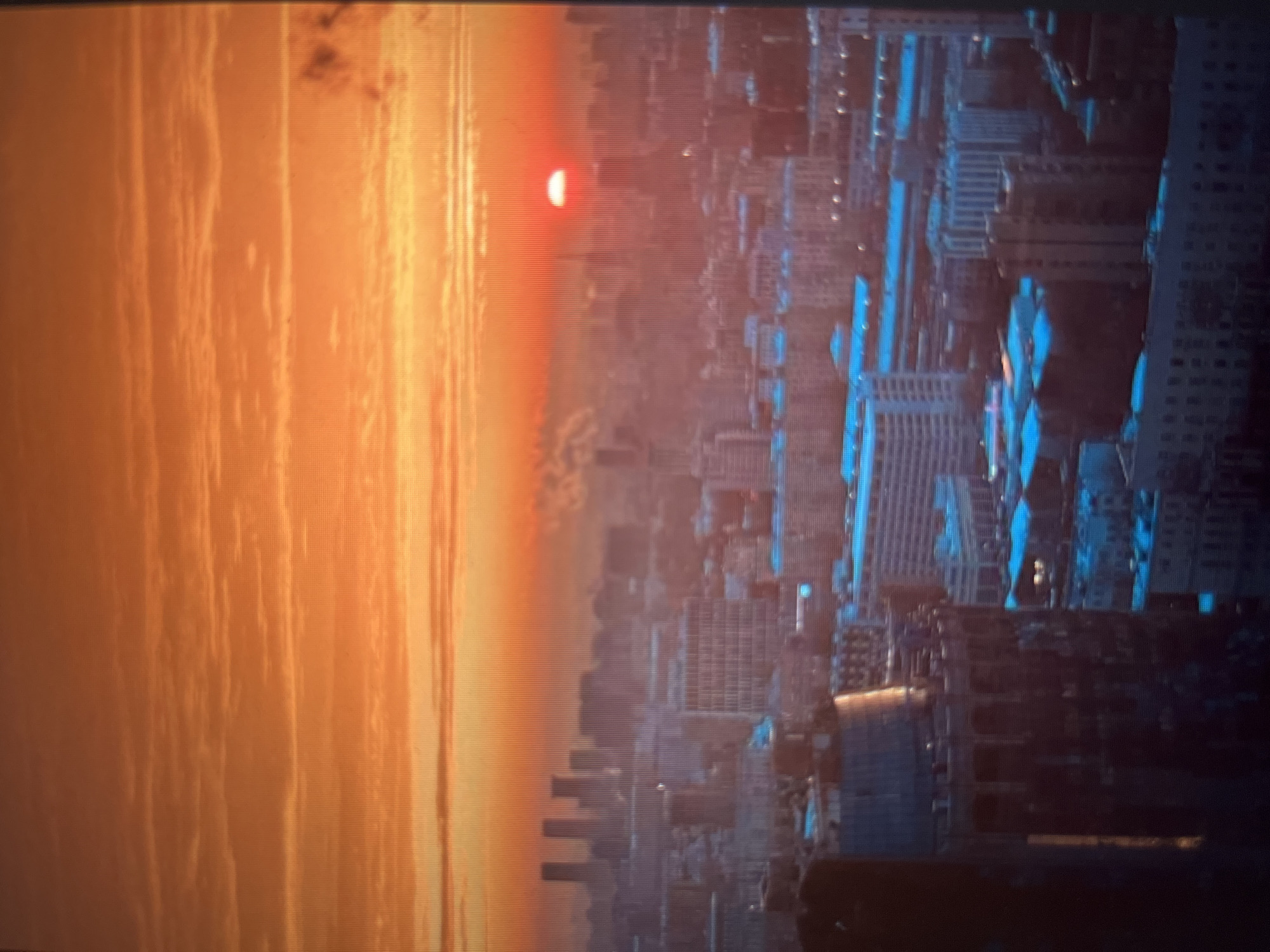 The image is a high-angle, long-shot photograph of a cityscape at sunset. The sky is a vibrant gradient of orange and yellow, with wispy clouds. The sun, a bright red orb, is partially visible on the horizon, casting a warm glow over the city. The city itself is a sprawling metropolis with numerous buildings of varying heights and architectural styles. In the foreground, a large, dark, cylindrical structure with arched openings dominates the left side of the frame. Further into the city, buildings appear to be illuminated with a blue light, possibly indicating artificial lighting or reflections. Some taller buildings, possibly skyscrapers, are visible in the distance, forming a more distinct skyline. The overall impression is one of urban density bathed in the dramatic light of a setting sun, with hints of activity suggested by the illuminated windows.