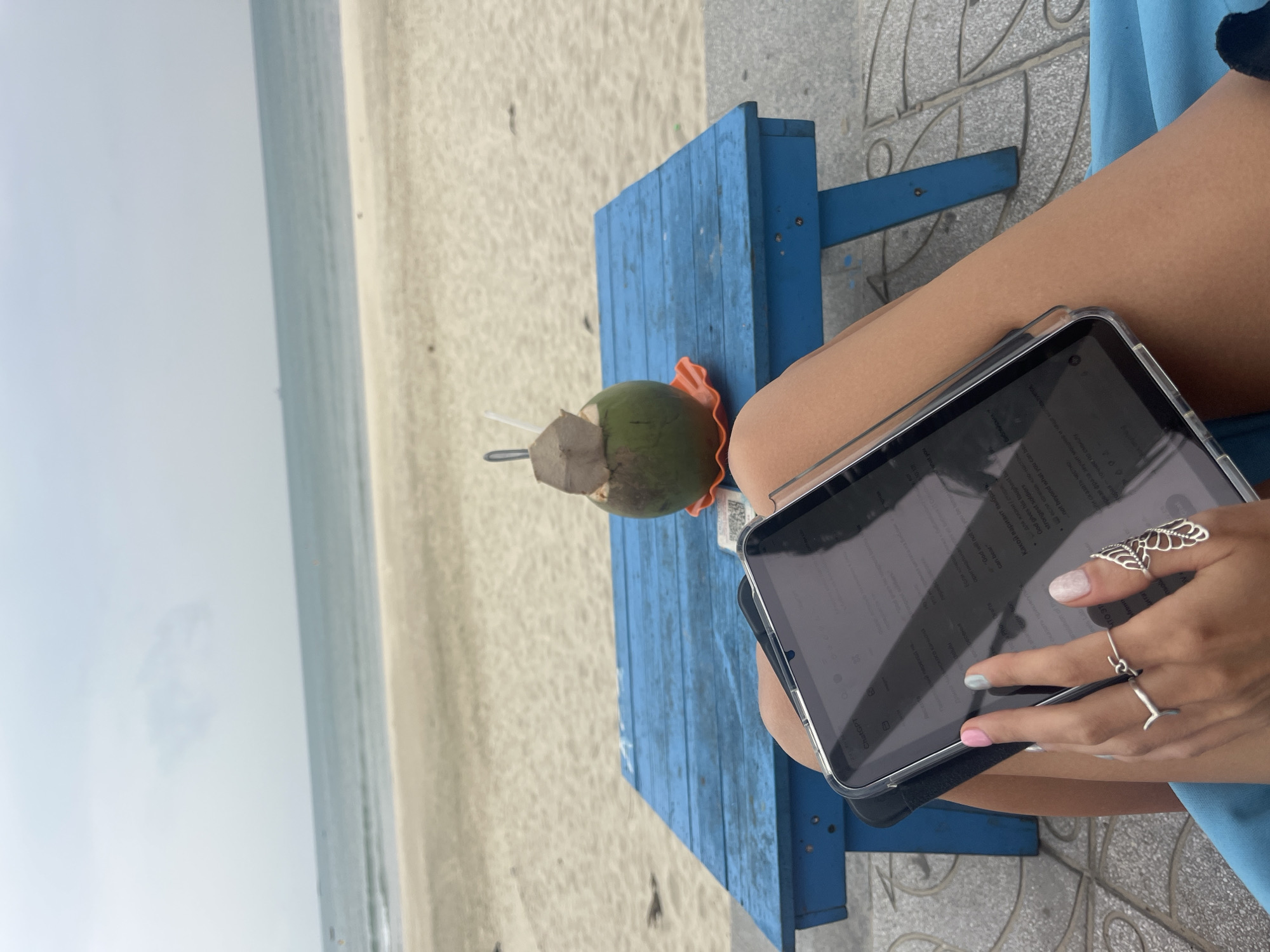 A person is seated by a beach in Đà Nẵng, Vietnam, holding a tablet and using it with their fingers. They are seated on a blue painted wooden bench. Beside them on the bench is a young coconut, with a straw and piece cut from the coconut shell. The beach and the ocean are visible in the background. The weather appears overcast. The person is wearing at least two rings on their hand.