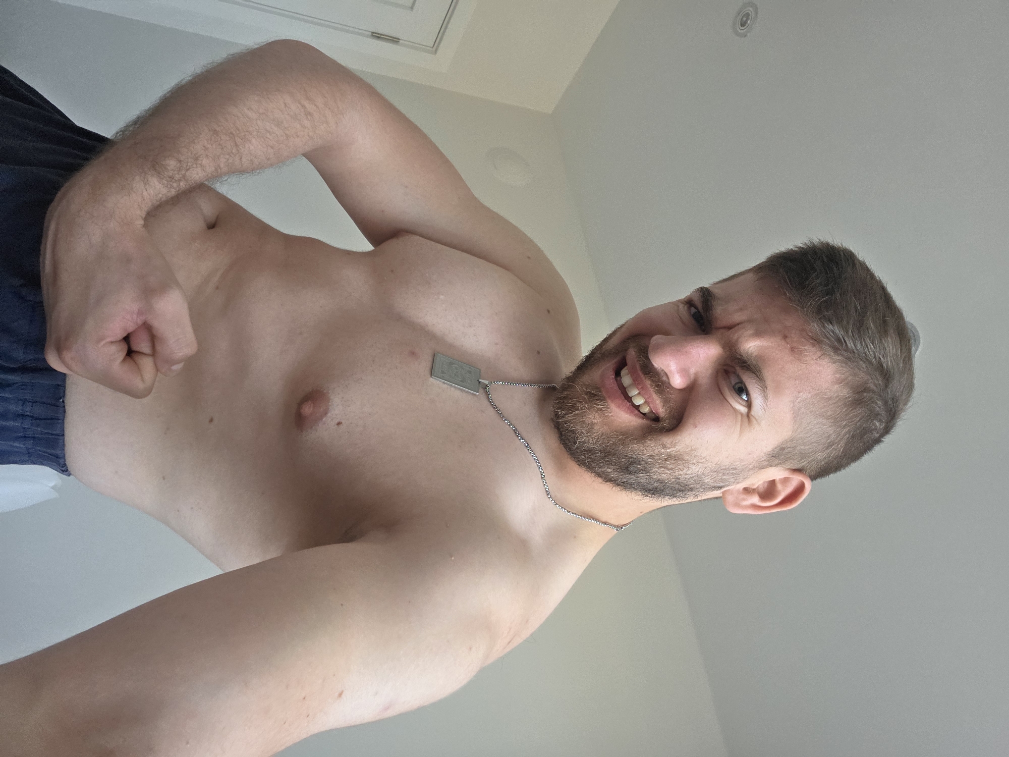 A shirtless man with a beard and short hair is flexing his right bicep. He is wearing blue pants and a silver dog tag necklace. The background consists of a white wall and a white door. The location is not discernible from the image.