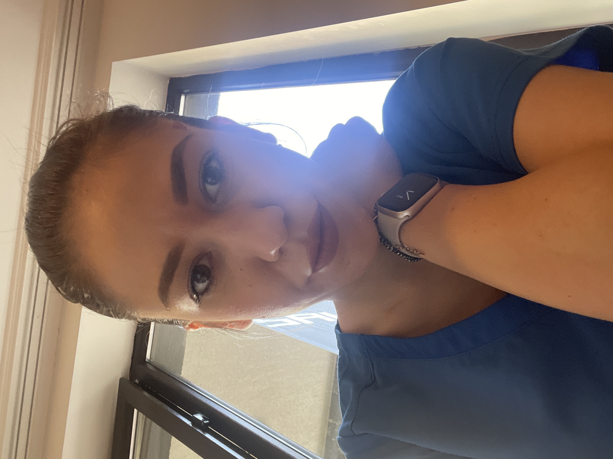 A close-up portrait features a young woman looking directly at the camera with a soft, gentle smile. Her dark hair is pulled back, and she has well-defined eyebrows and light makeup. She is wearing a royal blue top, possibly a scrub uniform or a simple t-shirt.
On her left wrist, she wears a modern smartwatch with a silver or rose gold case and a textured grey or black band. A small, dark-colored beaded bracelet is also visible just above the watch. Her left hand rests casually near her neck/collarbone.
The background is dominated by a large window with dark frames, which emits a very bright, almost overexposed light, suggesting it is daytime. This strong backlighting creates a subtle halo effect around her head and shoulders. To the left of the window, a light-colored wall is visible. The smartwatch screen displays abstract symbols and arrow-like icons, but no discernible text or numbers. The setting is indoors, with the context indicating a location in Sofia, Bulgaria.