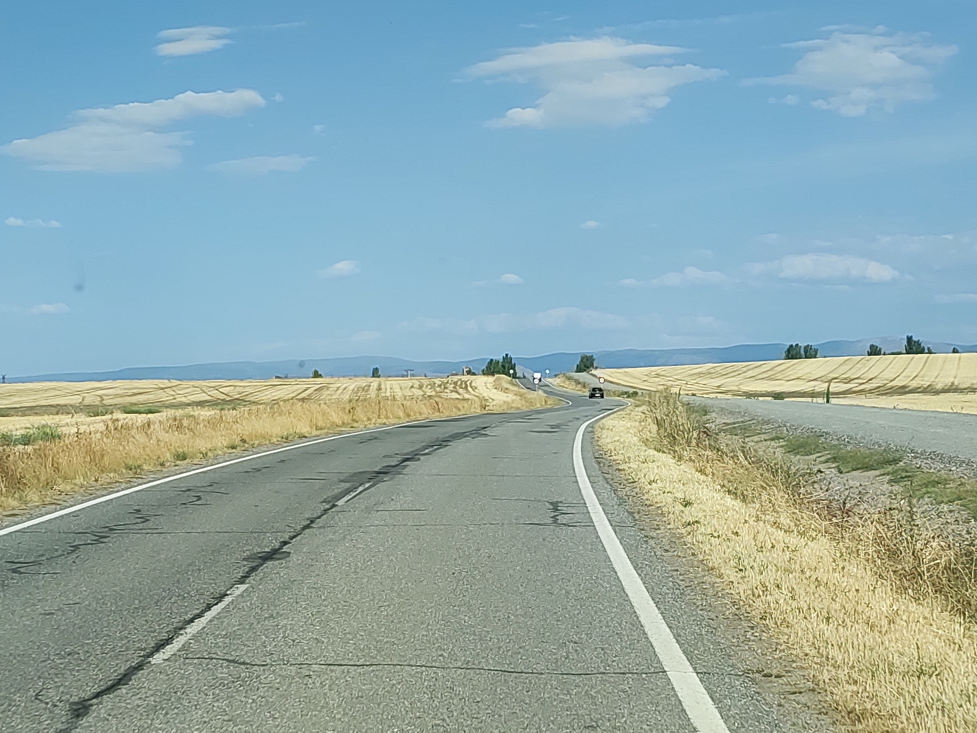 The image depicts a two-lane asphalt road curving through a rural landscape under a clear blue sky with scattered white clouds. The road is flanked by dry, golden fields of harvested crops, suggesting a late summer or early autumn setting. The fields exhibit distinct patterns of cultivation, with visible tracks from agricultural machinery. In the distance, a hazy line of hills or mountains forms the horizon. A single dark-colored car is visible on the right lane of the road, moving away from the viewer. The scene appears to be located in the vicinity of Abades, Spain, known for its agricultural plains and rural character. The overall impression is one of a quiet, open country road.