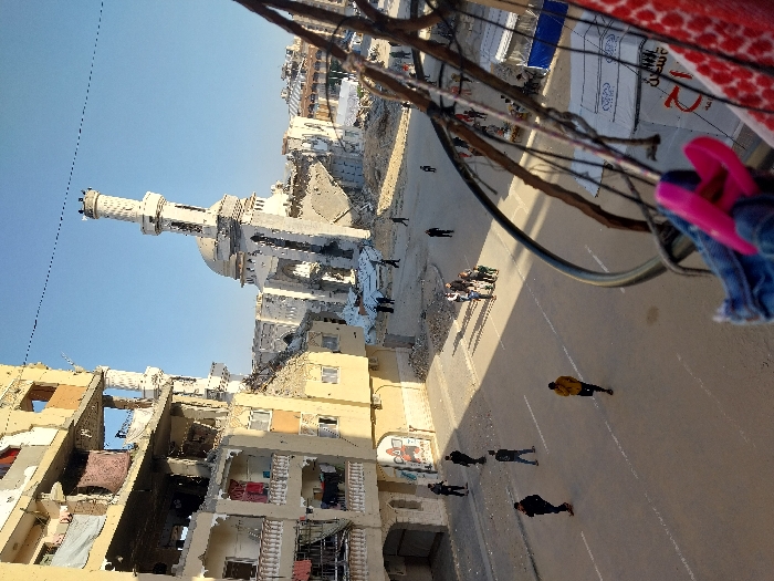 The image depicts an urban scene in Khan Yunis, Palestinian Territory, characterized by extensive structural damage under a clear blue sky.
In the mid-ground, a multi-story building, likely residential, is severely damaged on the left, with large sections of its facade and internal structures missing, exposing several floors. Laundry hangs from damaged balconies. Adjacent to it, towards the center-right, stands the heavily damaged Great Mosque of Khan Yunis (Al-Omari Mosque). Its distinctive dome is visible, and its tall minaret is partially collapsed and significantly tilted to the right. Surrounding the mosque are piles of debris and rubble. Further in the background, more damaged buildings and possibly makeshift shelters are faintly discernible.
The foreground shows a paved street with white markings. Approximately 15-20 individuals, including adults and several children, are present on the street and near the damaged structures. Some are walking individually, while a group of approximately five people, including children, walks together towards the right mid-ground. One individual in a yellow jacket walks alone in the center of the street. People appear to be moving through or observing the devastated environment.
From the perspective of the camera, elements of a balcony railing or metal frame are visible in the immediate foreground on the right, with a piece of blue denim fabric and pink flip-flops hanging from it, alongside a red and white patterned fabric. The scene is illuminated by direct sunlight, casting shadows.