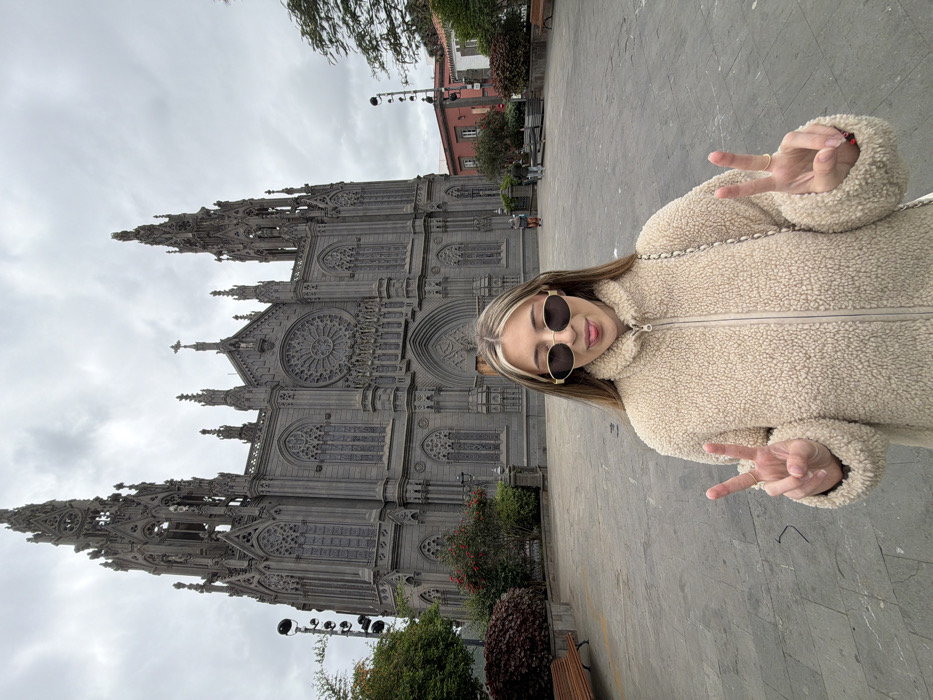 An outdoor, daytime scene under an overcast sky depicts a female subject standing in the foreground of a paved plaza, with the Parroquia de San Juan Bautista (Church of San Juan Bautista) in Arucas, Spain, dominating the background. The female, with light skin and long, highlighted hair, wears dark oval sunglasses with gold frames and a light beige fleece jacket. She faces the camera, extending her tongue slightly and displaying "peace signs" with both hands. A thin chain, possibly from a shoulder bag, is visible over her right shoulder.
The church, a large Gothic Revival structure, is constructed from dark grey volcanic stone, featuring two tall spires, a prominent central rose window, numerous arched windows, and intricate decorative elements. The plaza surface consists of light grey stone tiles. To the left, dark red and green bushes, a wooden park bench, and a tall black streetlight pole are present. To the right, further vegetation, another wooden bench, and a two-story building with reddish-orange walls are visible. Multiple indistinct figures are scattered in the mid-ground of the plaza, near the church's entrance.