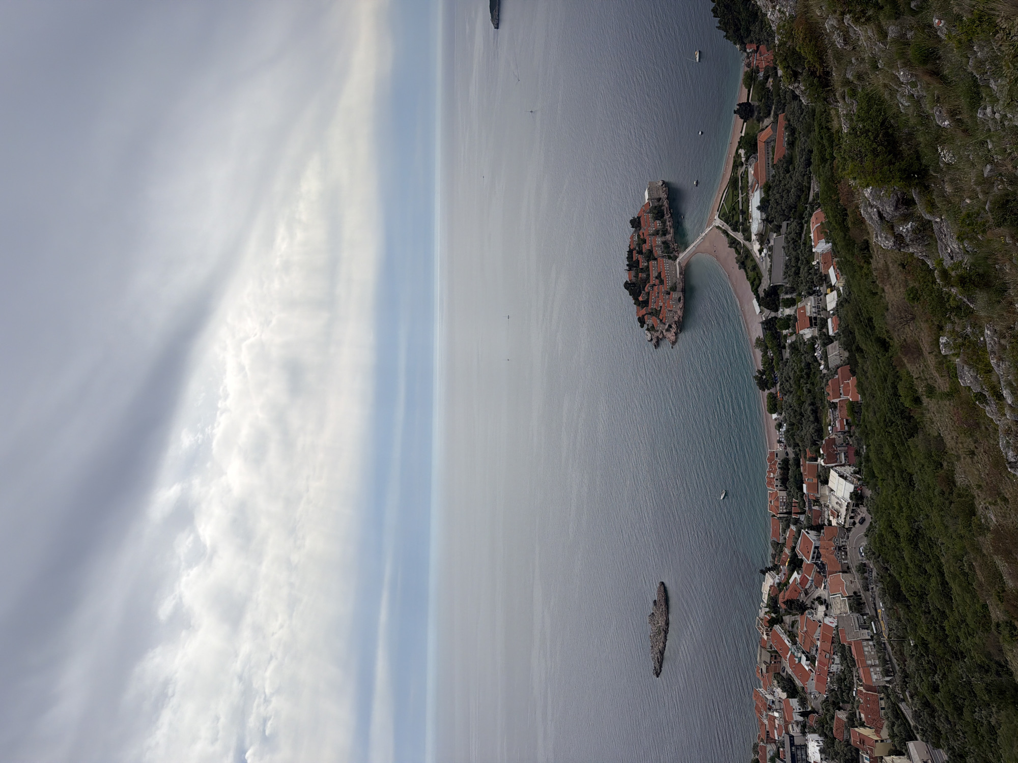 The image shows an aerial view of Sveti Stefan, Montenegro, with the island resort connected to the mainland by a narrow isthmus. The Adriatic Sea surrounds the island. The coastline features buildings with red-tiled roofs. Clouds are visible in the sky. A mountainous terrain is on the right. Another small island is visible in the sea.