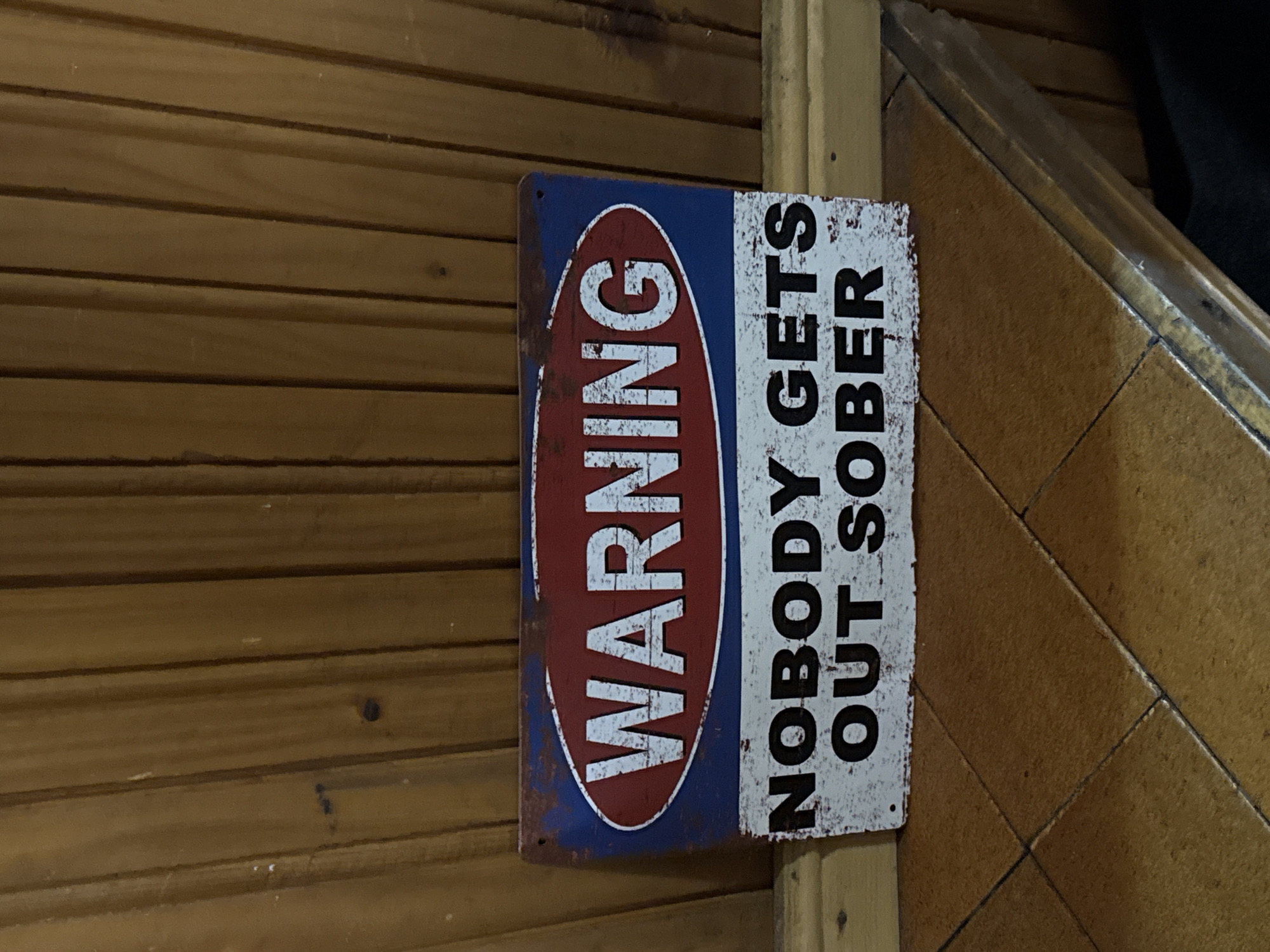 The image shows a weathered, rectangular metal sign mounted on a wood-paneled wall. The sign features the word "WARNING" vertically aligned in a red oval against a blue background on the left side. To the right, in black block letters against a white background, is the phrase "NOBODY GETS OUT SOBER." The sign exhibits signs of rust and wear. The surrounding environment consists of wooden paneling to the left and a vertical light wood trim piece to the right. The presence of tiled surfaces further to the right suggests the sign is indoors. This image doesn't indicate the location is in Lilkovo, Bulgaria.