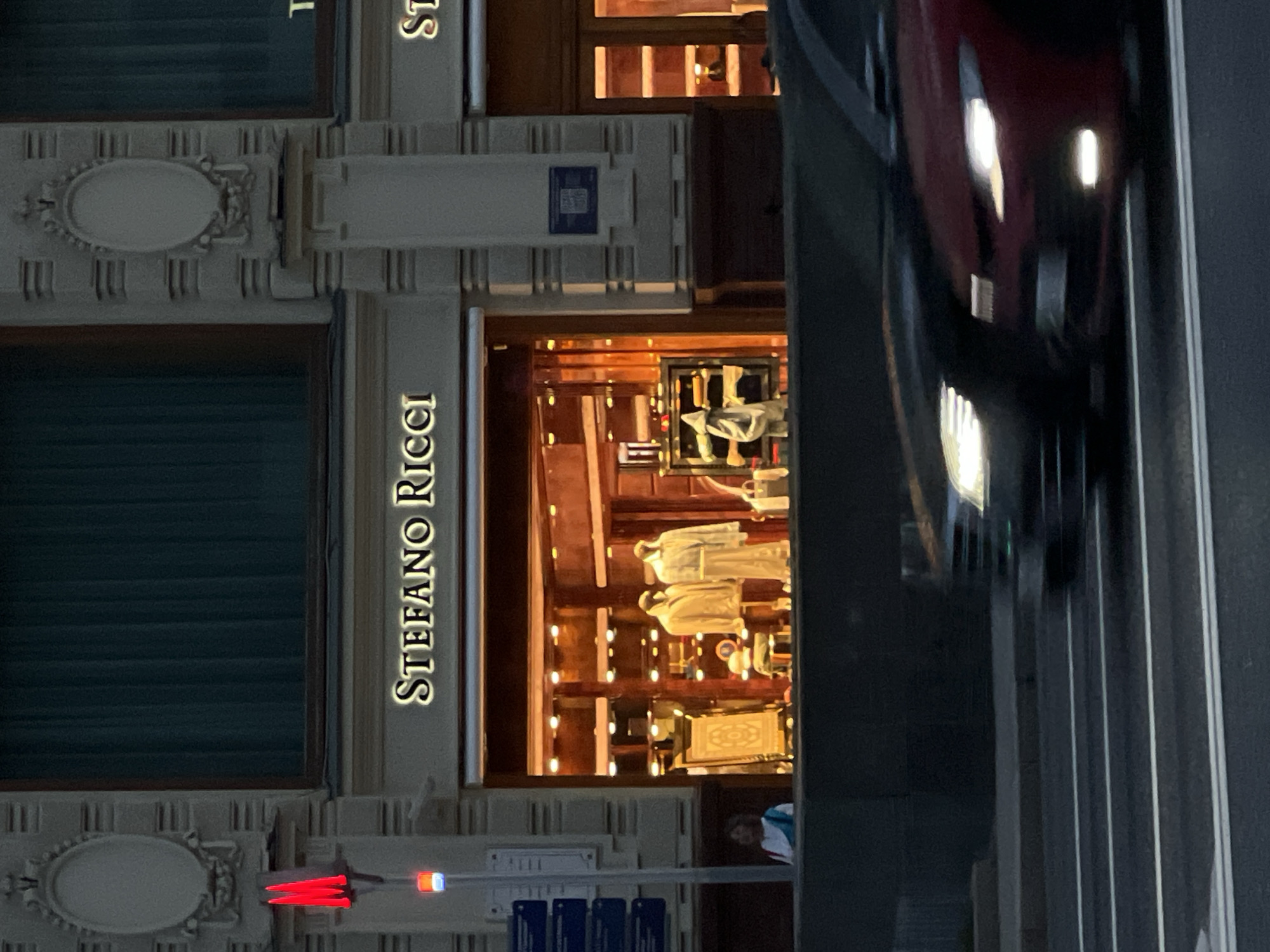 The image depicts the storefront of a Stefano Ricci boutique. The store's name is vertically oriented on the facade. Through the store's windows, mannequins are visible. Outside the store, traffic moves along a road, with a red car's headlights captured in motion. Street furniture such as traffic lights is also visible. Based on the architectural style, the storefront is likely located in Moscow, Russia.