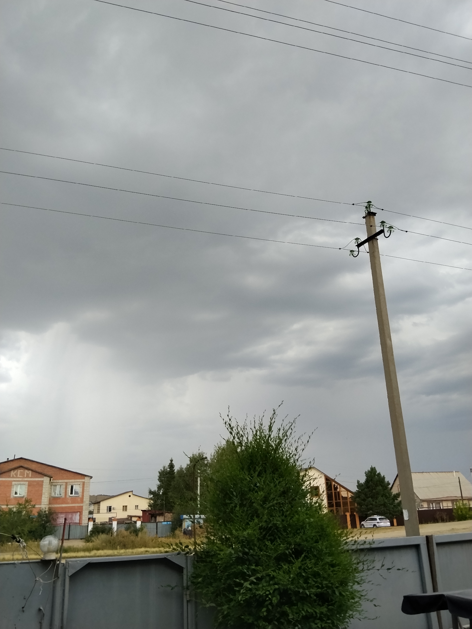 The image depicts a cloudy sky over a residential area with several houses and a prominent power pole. On the left, a brick building with "KCM" visible on its facade is present. In the background, various houses with different architectural styles are scattered. A white car is parked in front of one of the houses on the right side of the image. A dense green bush occupies the foreground on the right, and a grey metal fence runs across the bottom of the frame. The overall scene suggests an overcast day in a suburban or rural setting. The location is identified as Kokshetau, Kazakhstan.