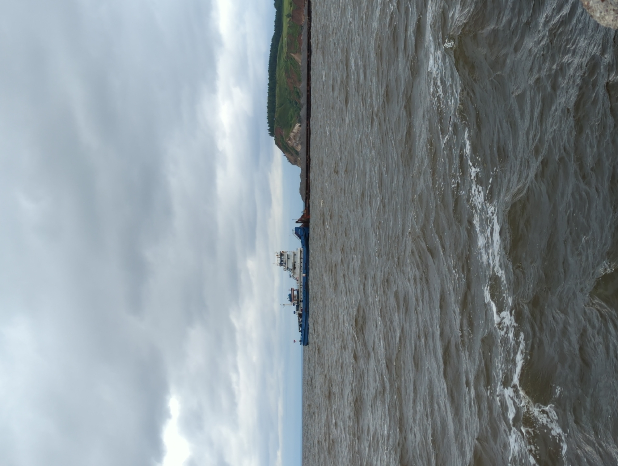 In Kamskoye Ustye, Russia, a wide body of water is depicted under a cloudy sky. The water is choppy with moderate waves and whitecaps. In the mid-ground, a blue and white vessel, appearing to be a tugboat, is pulling a long barge filled with gravel. The barge extends horizontally across the frame, its cargo piled high. In the background, a lush green cliff rises from the water's edge, topped with a dense line of dark green trees. The overall scene suggests a working day on the water, with the weather being overcast.