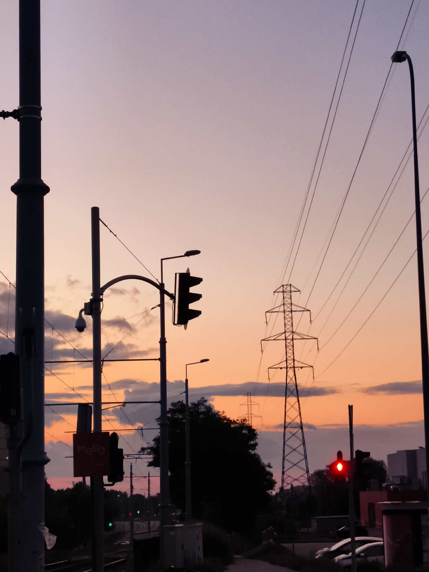 The image depicts a twilight scene in the city of Gdańsk, Poland, near the railway tracks. A tall, dark lamppost stands on the left, partially obscuring a large dark pole with a security camera. In the center, a traffic signal with a red light is illuminated, mounted on a curved pole alongside another dark pole. Further to the right, another lamppost is visible, and a large electrical pylon dominates the background. The sky displays a gradient of purple and orange hues, with scattered clouds. In the foreground, railway tracks are visible, and a dark red sign with white lettering reads "MeVo". In the distance, trees and buildings are silhouetted against the sky, with hints of other traffic lights showing green.